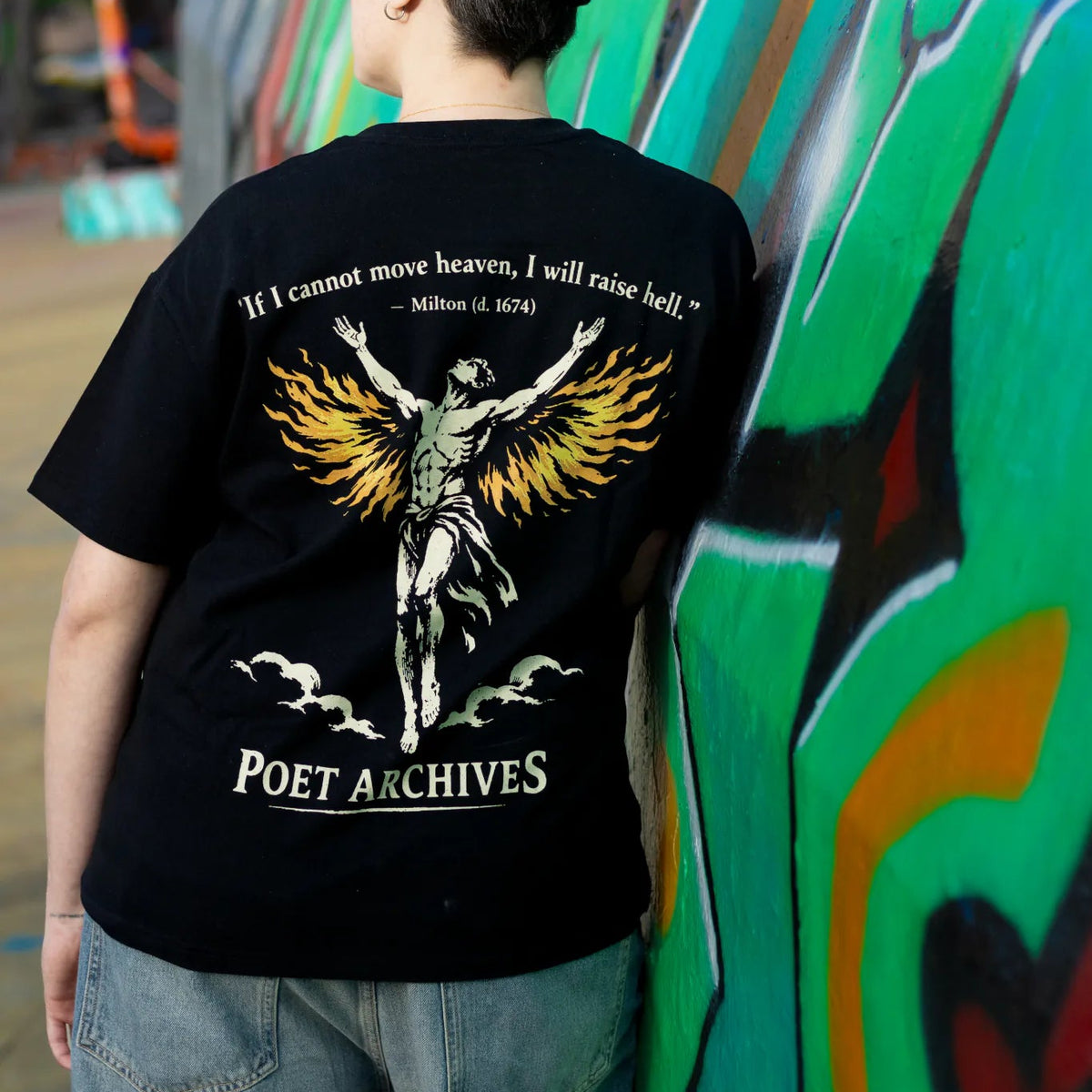 A person wearing a black graphic t-shirt with a detailed winged figure and text print on the back, standing in front of a colorful graffiti-covered wall.