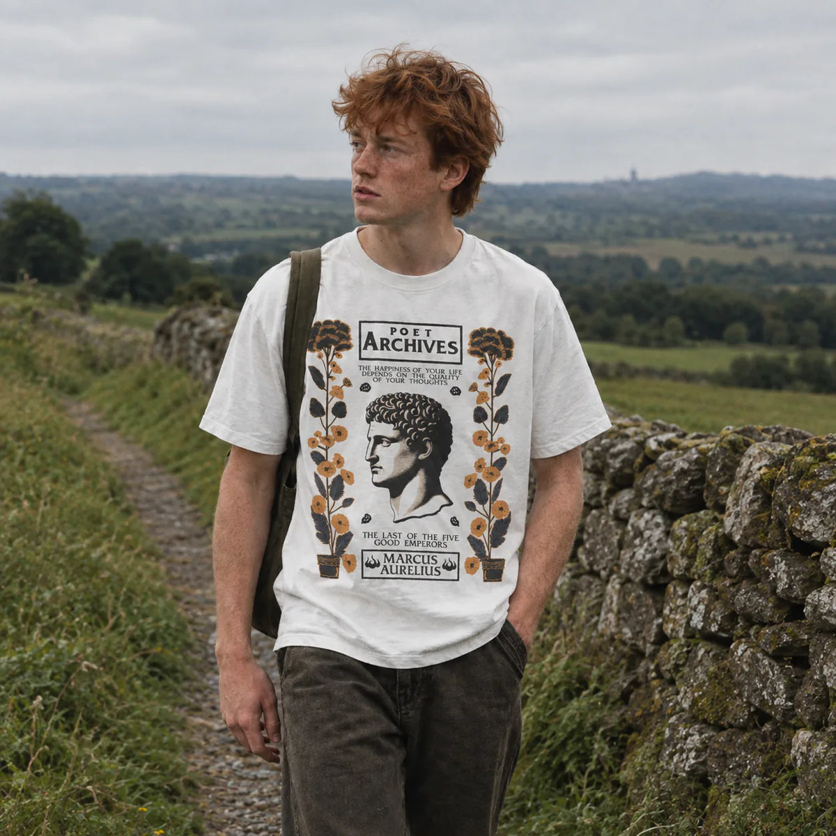 A man with red hair stands on a rural path wearing a white graphic T-shirt with a vintage-style poster print featuring a profile portrait and floral elements, along with dark trousers and a shoulder bag, set against a backdrop of green fields and a stone wall.