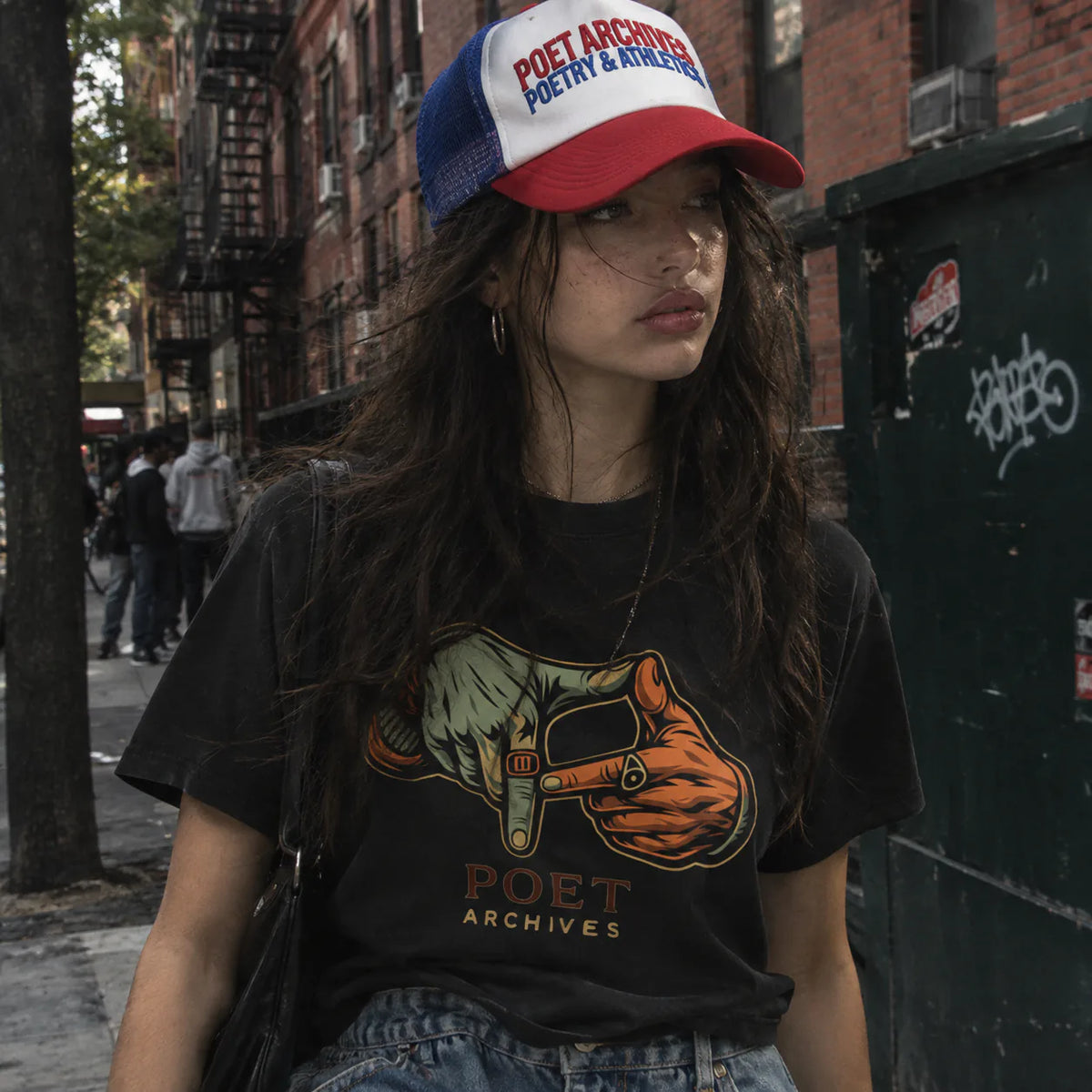 A woman wearing a red, white, and blue trucker hat with 'POET ARCHIVES' text and a black graphic t-shirt with a matching 'POET ARCHIVES' print, standing on a city street.