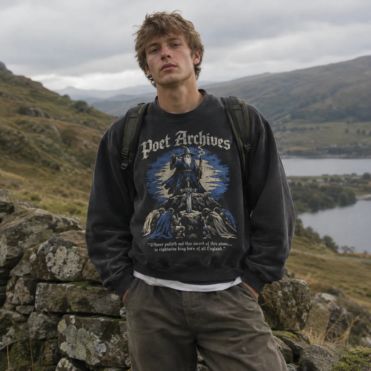 A young man standing outdoors in a mountain landscape wearing a dark charcoal graphic sweatshirt with 'Poet Archives' printed across the chest, olive pants, and a black backpack.