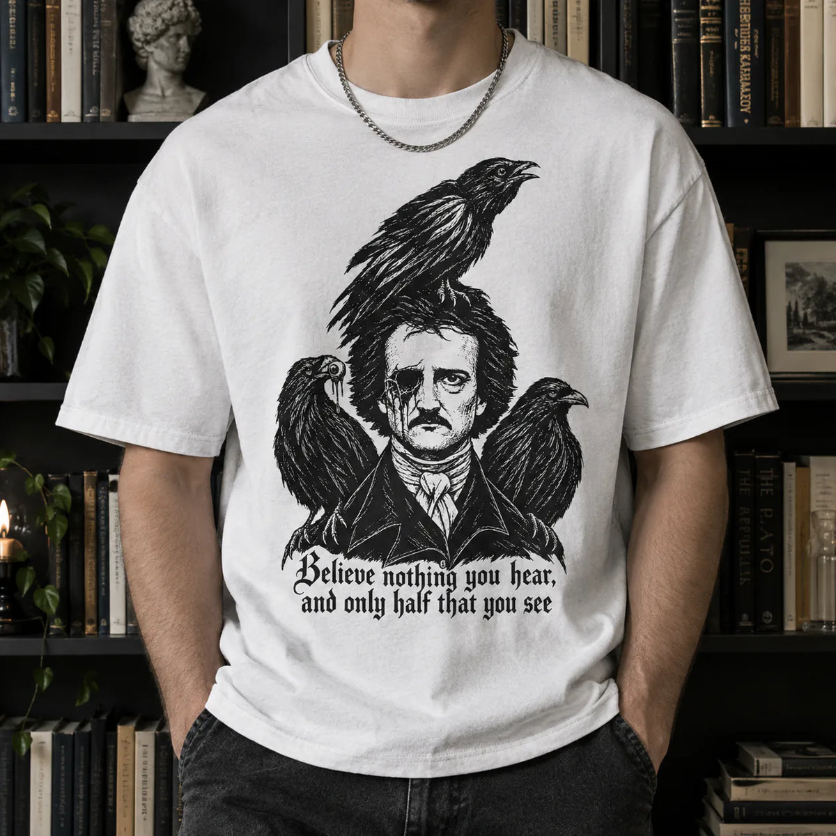 A man wearing a white graphic t-shirt featuring a black illustration of Edge from The Addams Family with crows and the quote 'Believe nothing you hear, and only half that you see'. The background shows dark bookshelves.