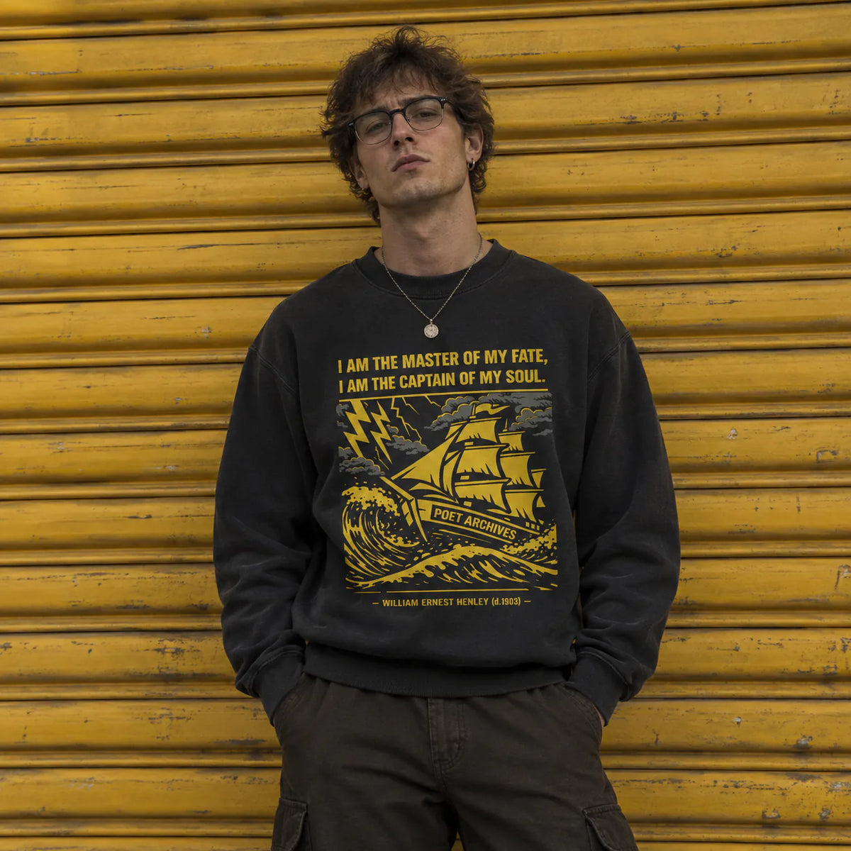 A man wearing a black crewneck sweatshirt with a yellow graphic print of a ship and a quote by William Kennedy O'Neal, standing in front of a yellow corrugated metal shutter.