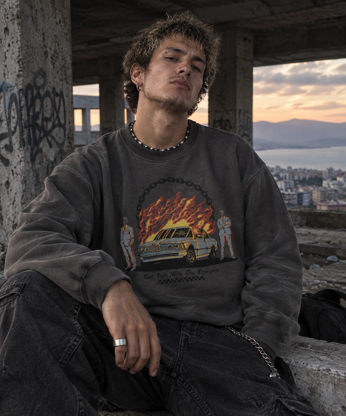 A man wearing a dark gray graphic sweatshirt and black jeans, sitting in a concrete urban setting at sunset.