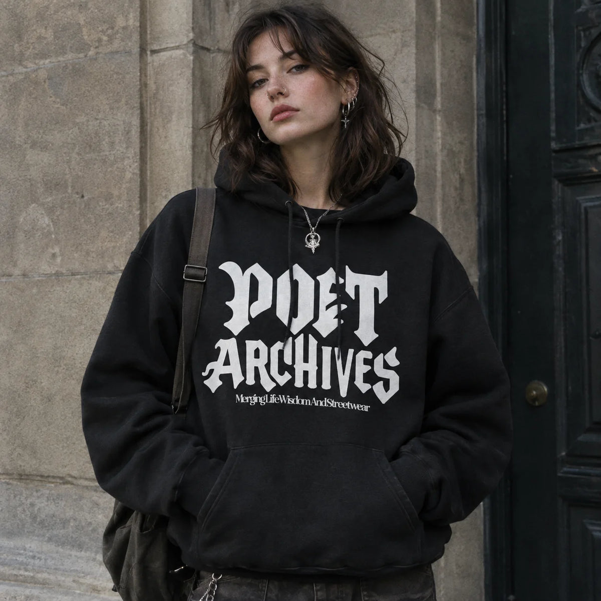 A woman wearing a black pullover hoodie with 'POET ARCHIVES' printed in a white gothic font on the front, standing against a stone wall and dark door.