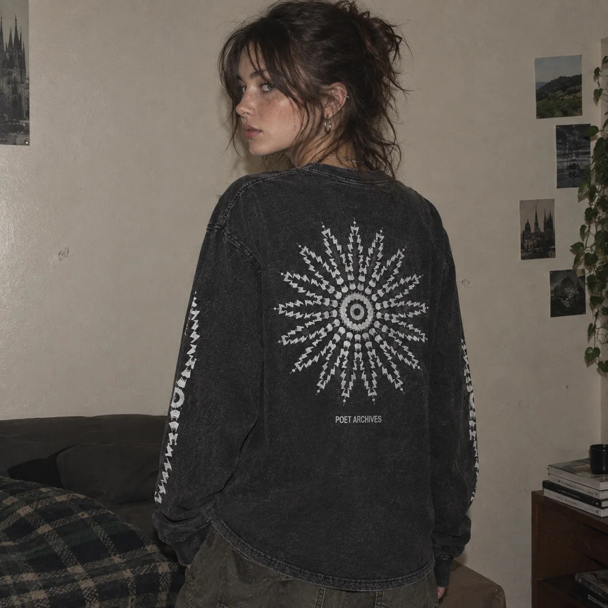 A person wearing a faded black washed long-sleeve shirt with a large white sunburst graphic and the text 'POET ARCHIVES' on the back, standing in a room with posters on the wall.