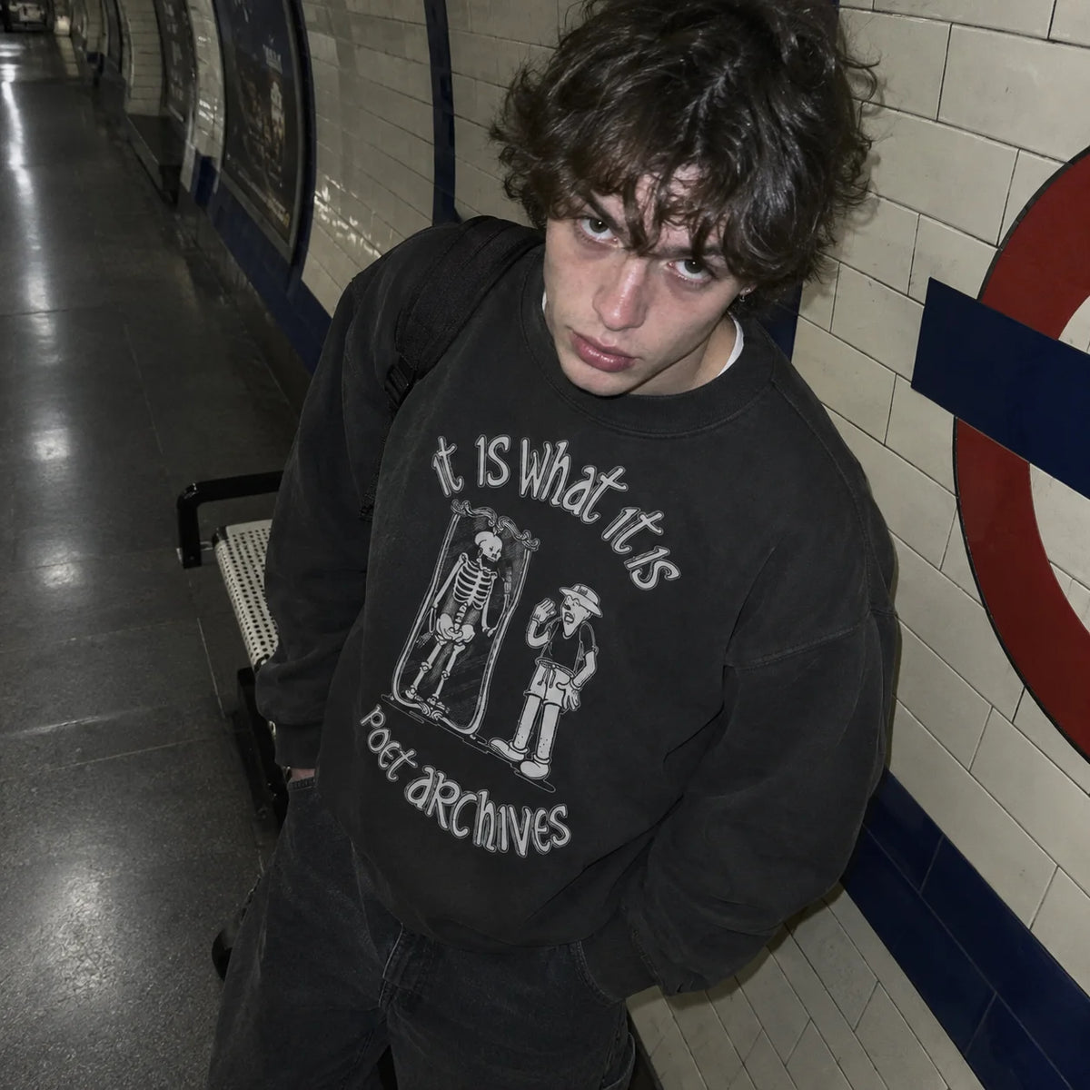 A person standing in a subway station wearing a black crewneck sweatshirt with a white graphic print that reads 'it IS what it IS poet archives' with two illustrated figures.