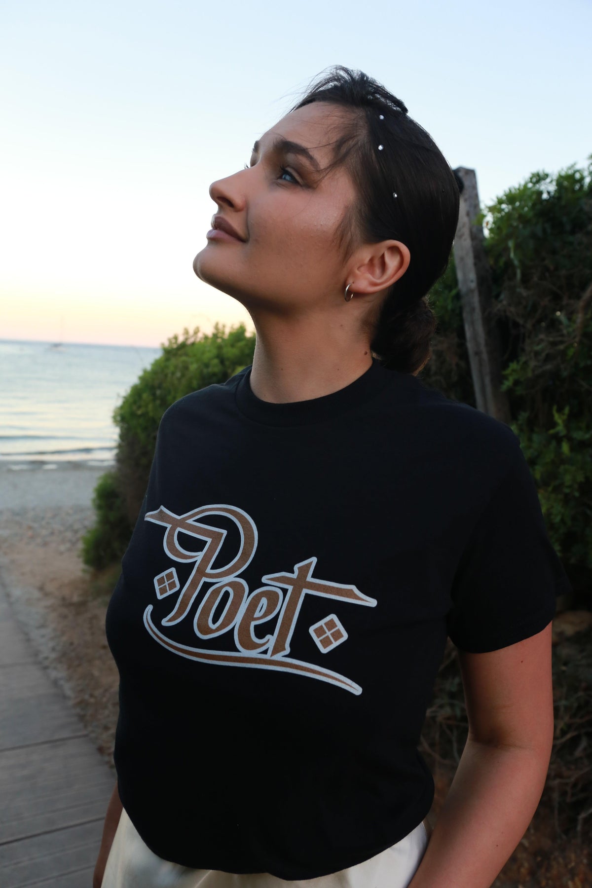 Woman wearing a black 'Poet' logo t-shirt on a coastal path at sunset