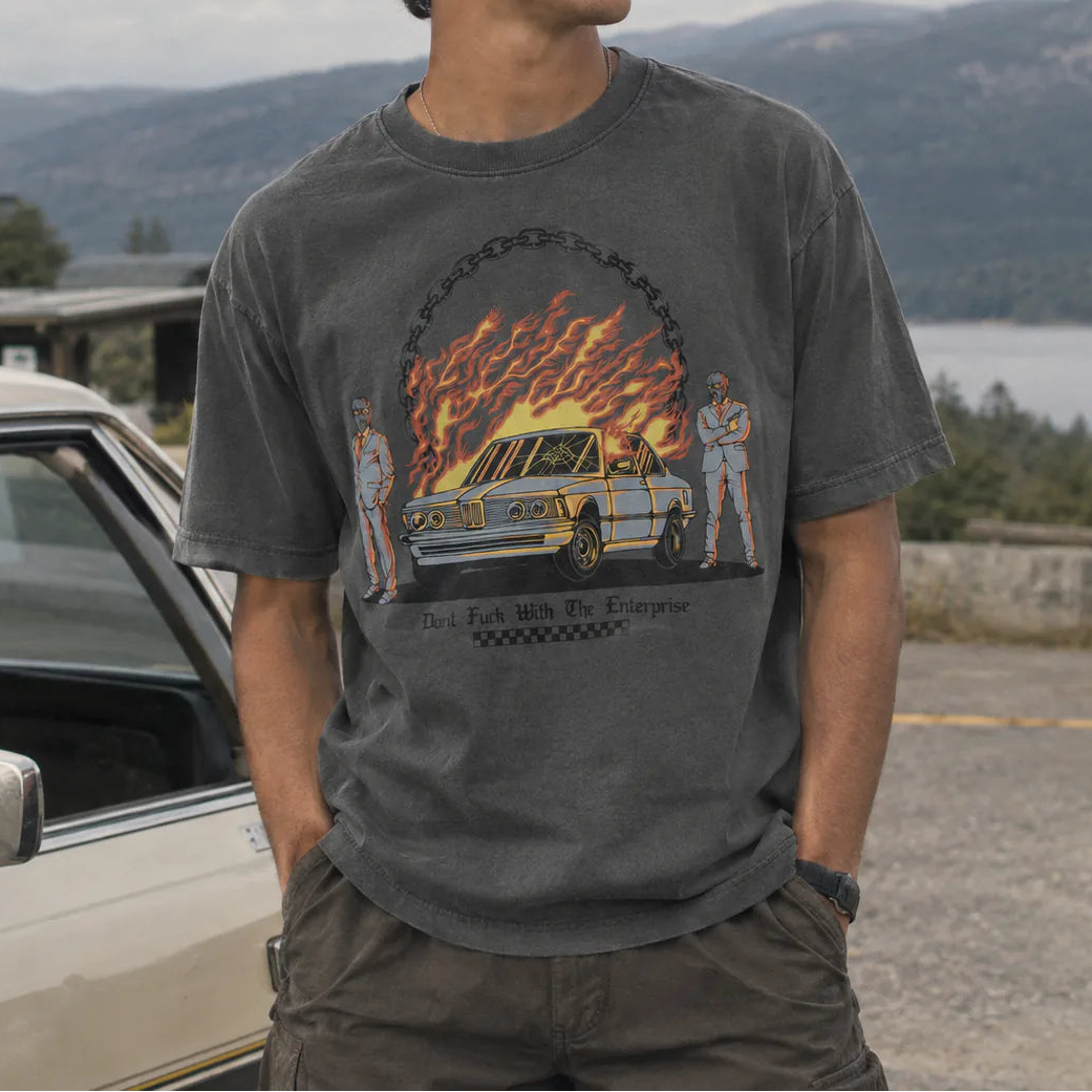 A man wearing a dark gray graphic t-shirt with a 'Don't Fuck With The Enterprise' car design, standing outdoors next to a vintage car with a mountain and lake background.
