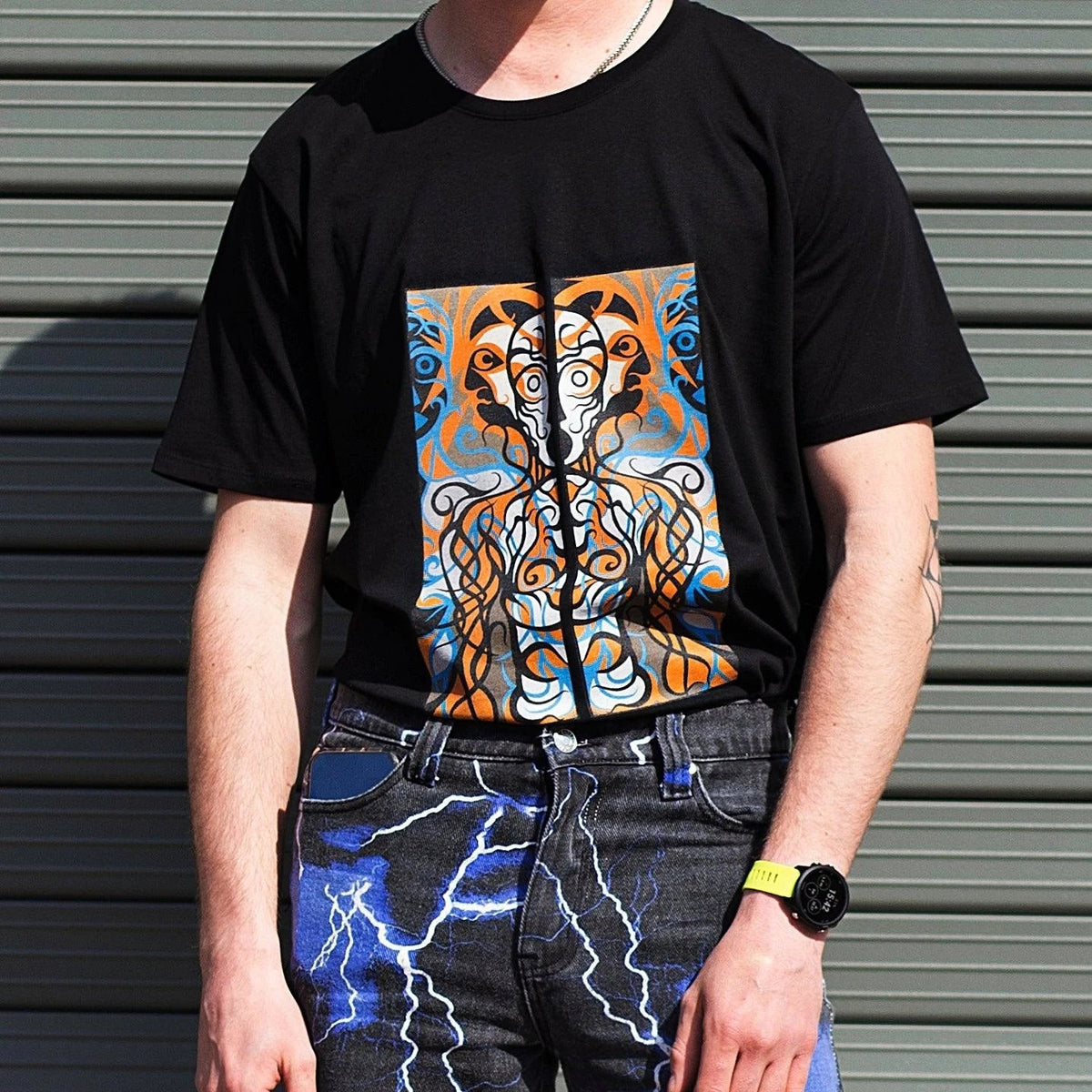 A person wearing a black short-sleeve t-shirt with a large square graphic print of a stylized face in orange, blue, and white, paired with dark jeans featuring a blue lightning pattern, standing in front of a gray metal shutter.