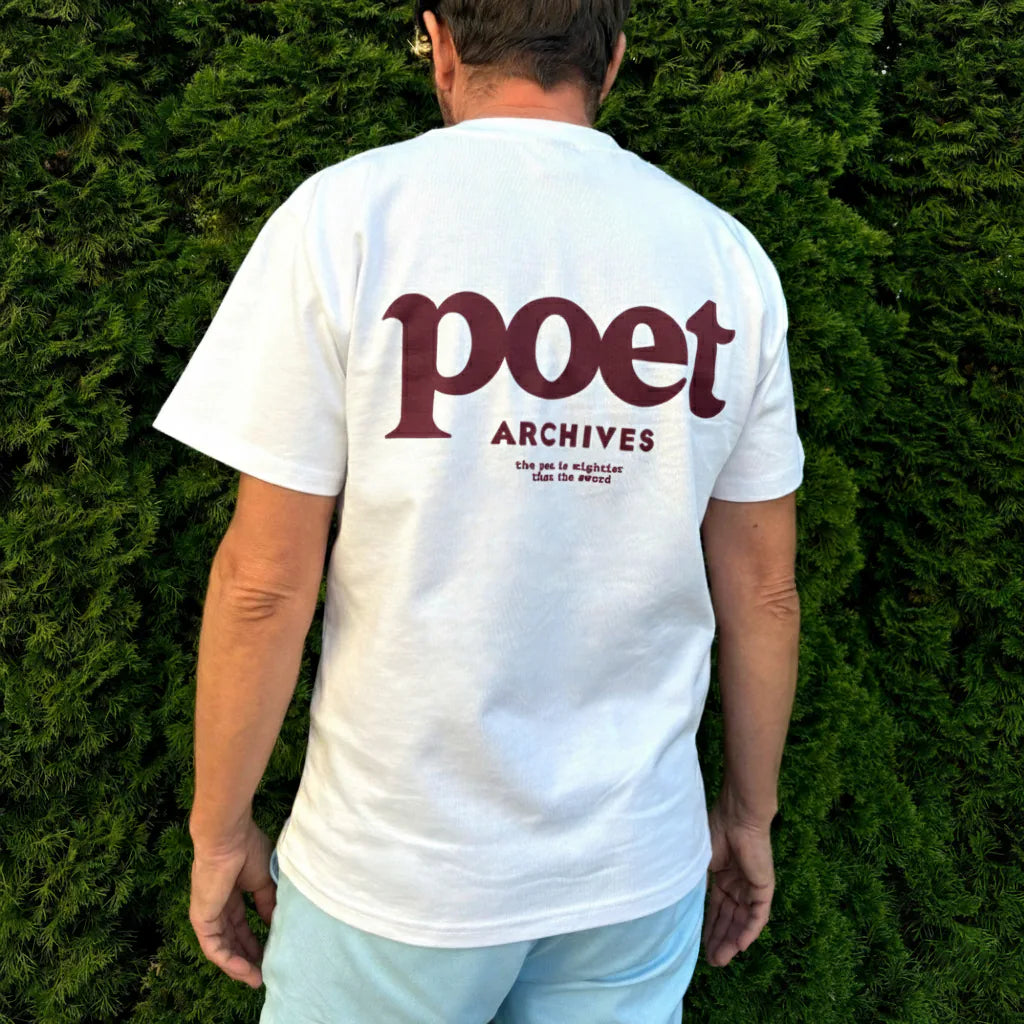 Person wearing a white t-shirt with 'poet ARCHIVES' text on the back, standing against a green hedge.