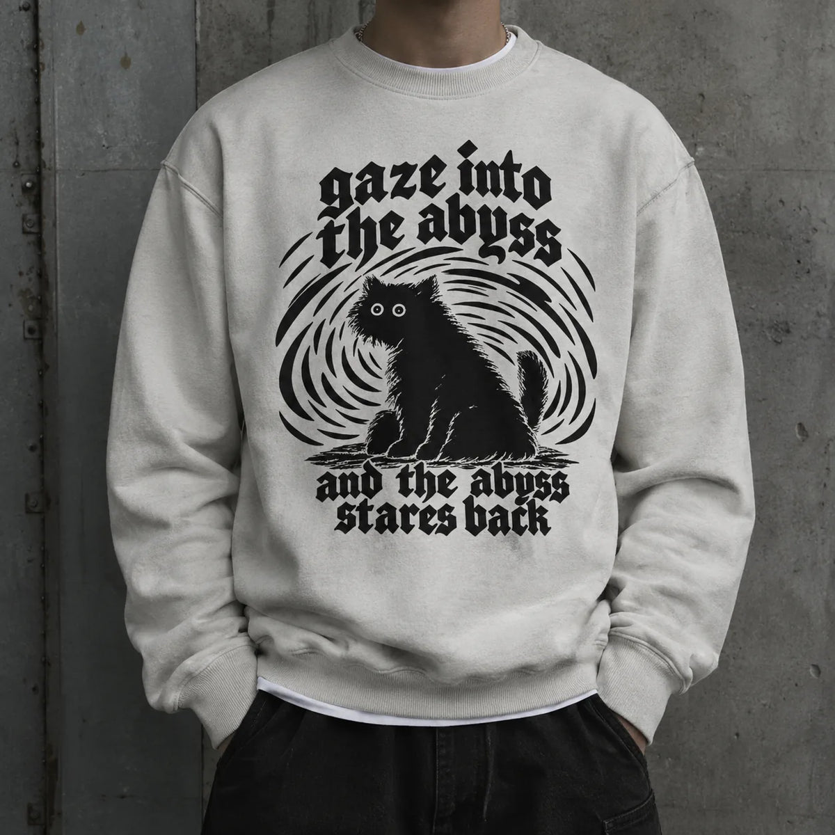 A person wearing a light gray crewneck sweatshirt with a black graphic of a cat and gothic text reading 'gaze into the abyss and the abyss stares back', posed against a textured gray concrete wall.