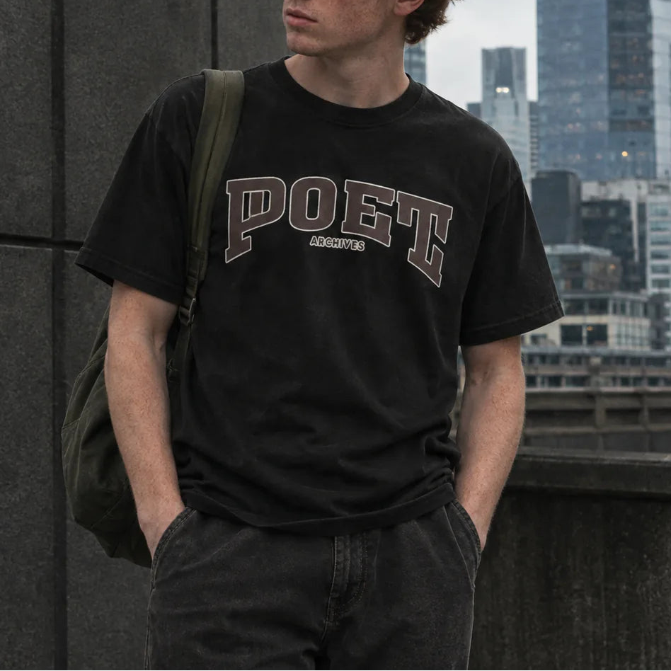 A young man with red hair stands outdoors wearing a black graphic t-shirt with 'POET' printed in arched varsity letters across the chest. The background shows a modern city skyline with skyscrapers under an overcast sky.