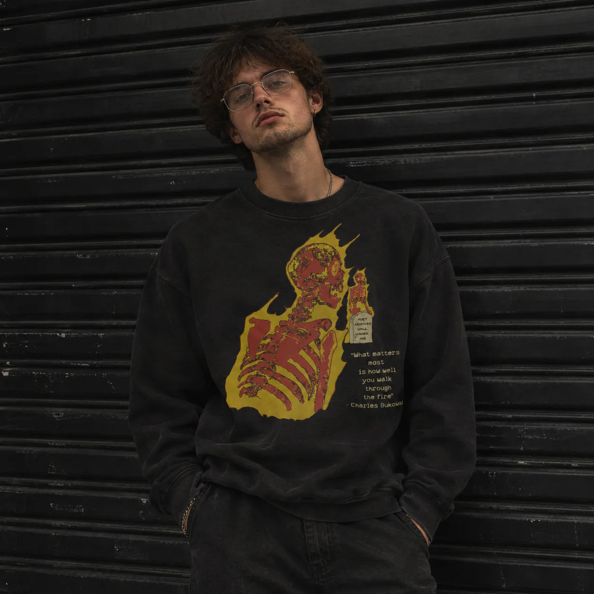 A man wearing a black crewneck sweatshirt with a red and yellow graphic of a skeleton and a bottle, standing in front of a black metal shutter.
