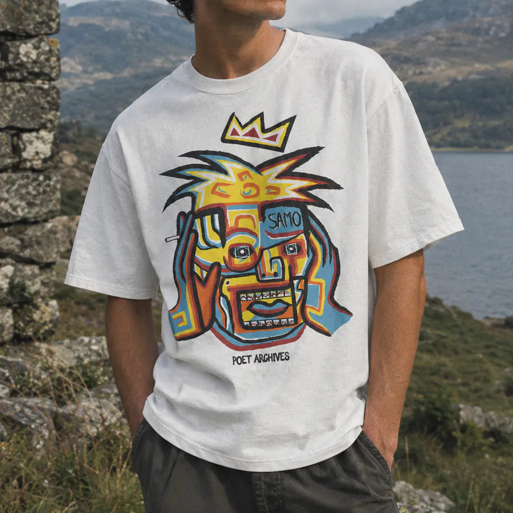 A man wearing a white graphic t-shirt with a colourful stylized face design and the text 'POET ARCHIVES' and 'SAMO'. He is standing outdoors in front of a stone wall with a lake and mountains in the background.