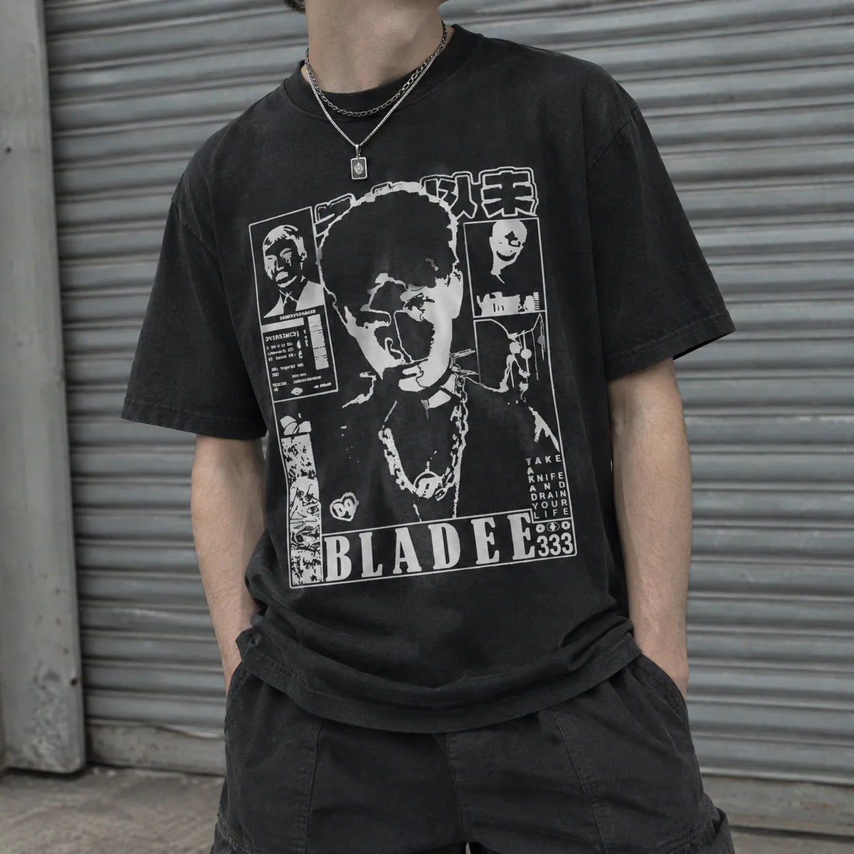 A person wearing a faded black graphic T-shirt with a large white and gray collage-style print featuring the word BLADEE and Japanese text, paired with dark shorts in front of a gray metal roll-up door.