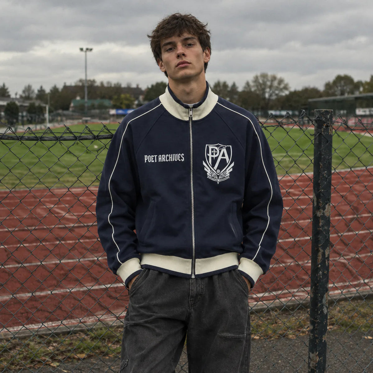A young man stands outdoors in front of a chain-link fence at an overcast track and field stadium, wearing a navy blue zip-up varsity-style jacket with cream ribbed trim and white embroidered graphics on the chest, paired with dark gray jeans.