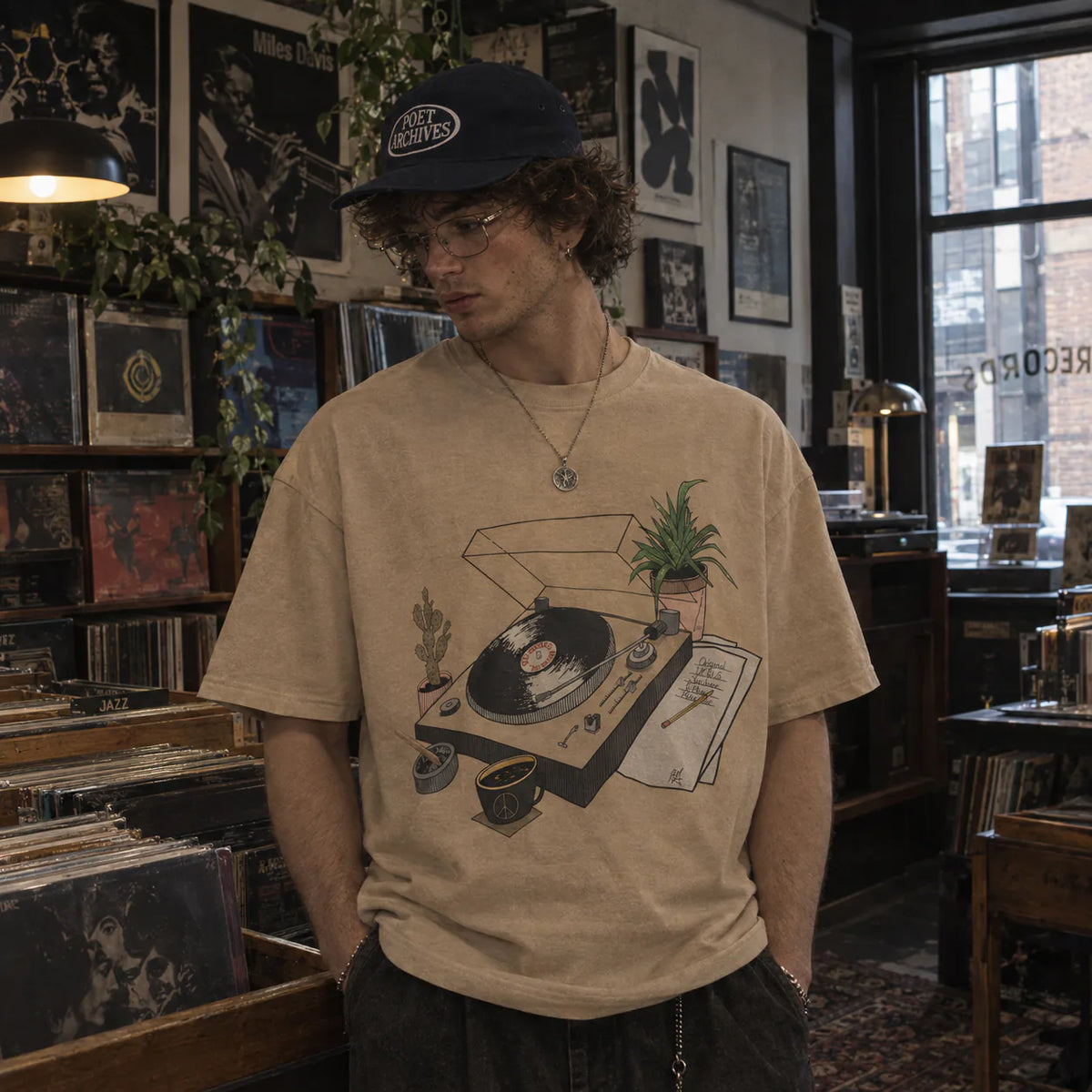 A man wearing a beige graphic t-shirt with a vinyl record and plant illustration, a dark navy baseball cap, and glasses, standing in a record store.