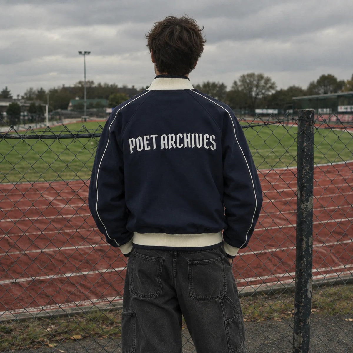 A person standing outdoors at a chain-link fence on an athletic field, viewed from behind, wearing a navy blue bomber jacket with white piping and 'POET ARCHIVES' printed in white across the back.