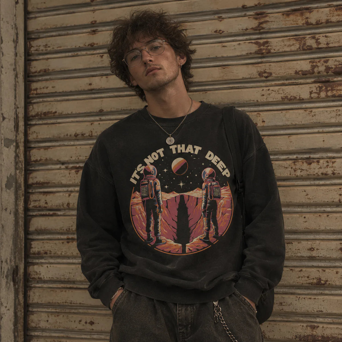 A man wearing a black graphic sweatshirt with a retro space-themed print and the text 'IT'S NOT THAT DEEP', standing in front of a weathered metal shutter.