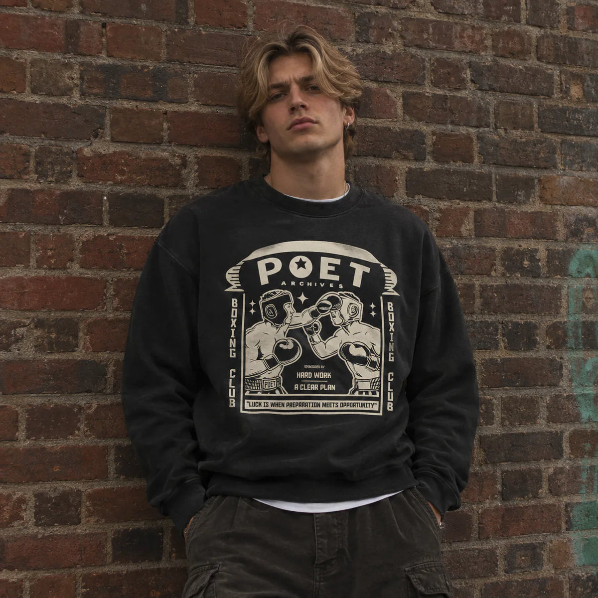 A young man with blonde hair stands against a red-brown brick wall wearing a black crewneck sweatshirt with a large white graphic print that reads 'POET ARCHIVES' and features boxing illustrations. He is also wearing dark cargo pants.