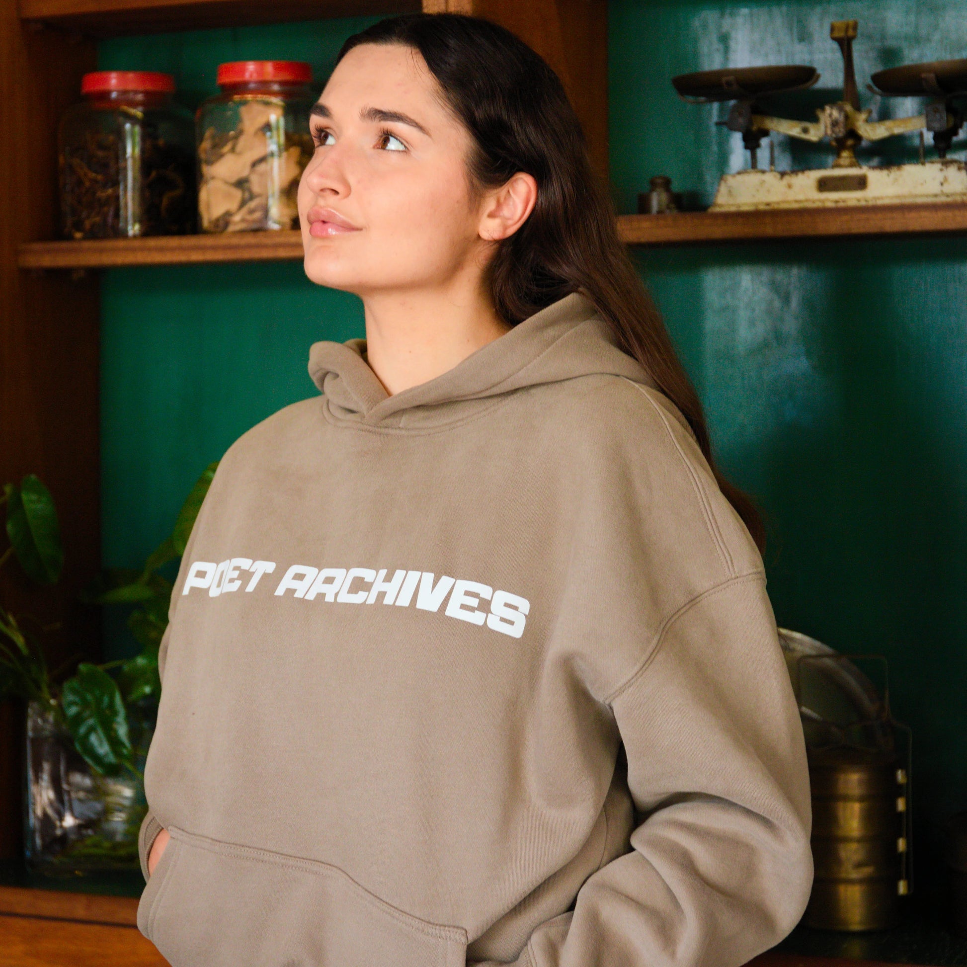 Person wearing a beige hoodie with 'PET ARCHIVES' text in a room with shelves and decor.