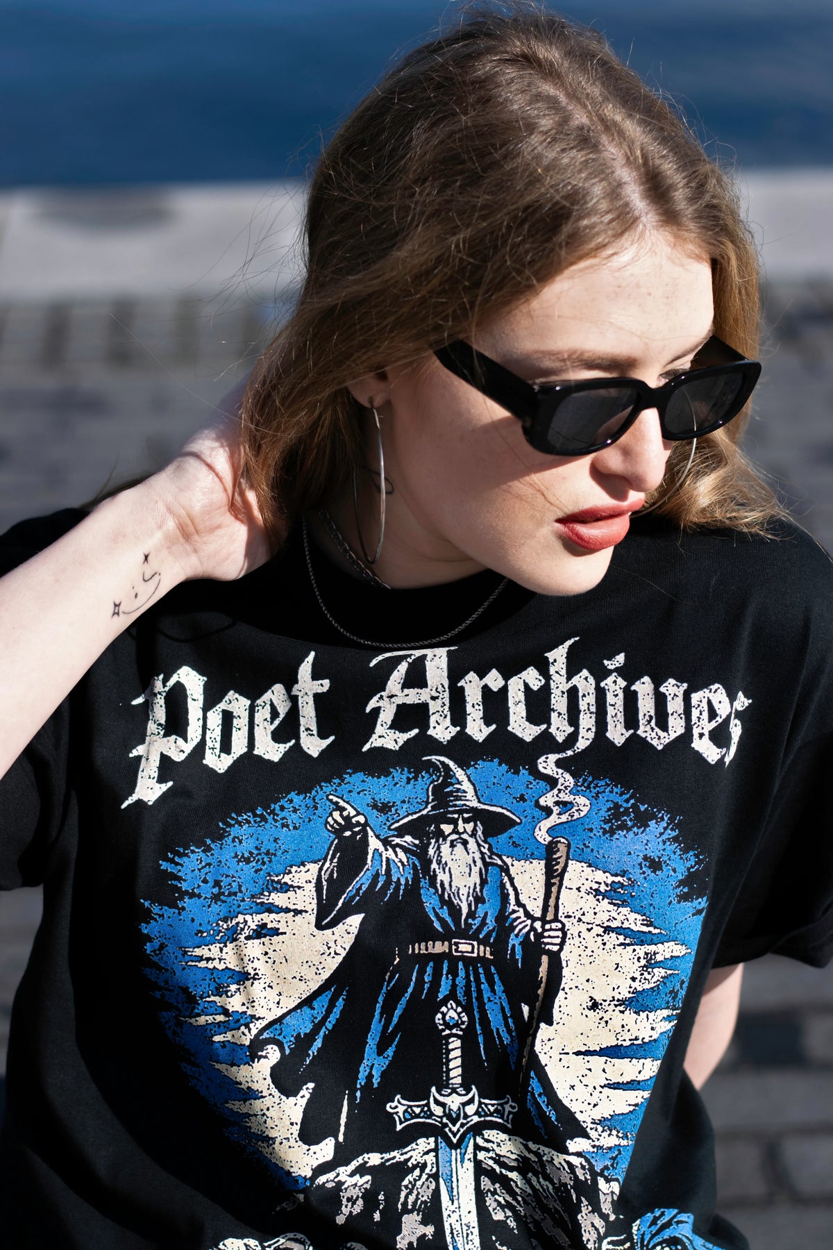 A woman wearing a black graphic t-shirt with 'Poet Archives' and a wizard illustration, photographed outdoors with a blurred stone wall and blue sky in the background.