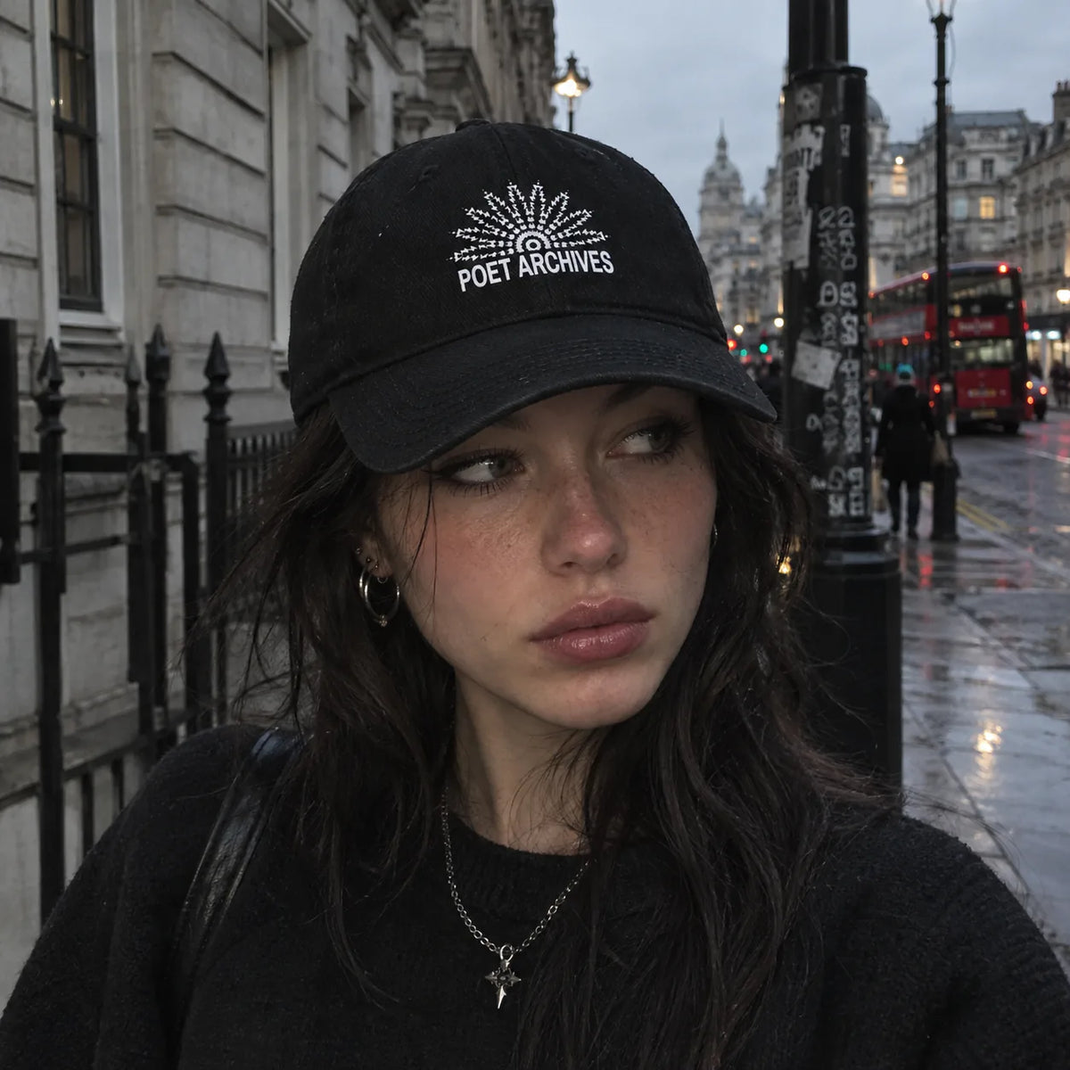 A woman wearing a black baseball cap with a white rhinestone 'Poet Archives' logo, photographed outdoors on a city street with a red double-decker bus in the background.