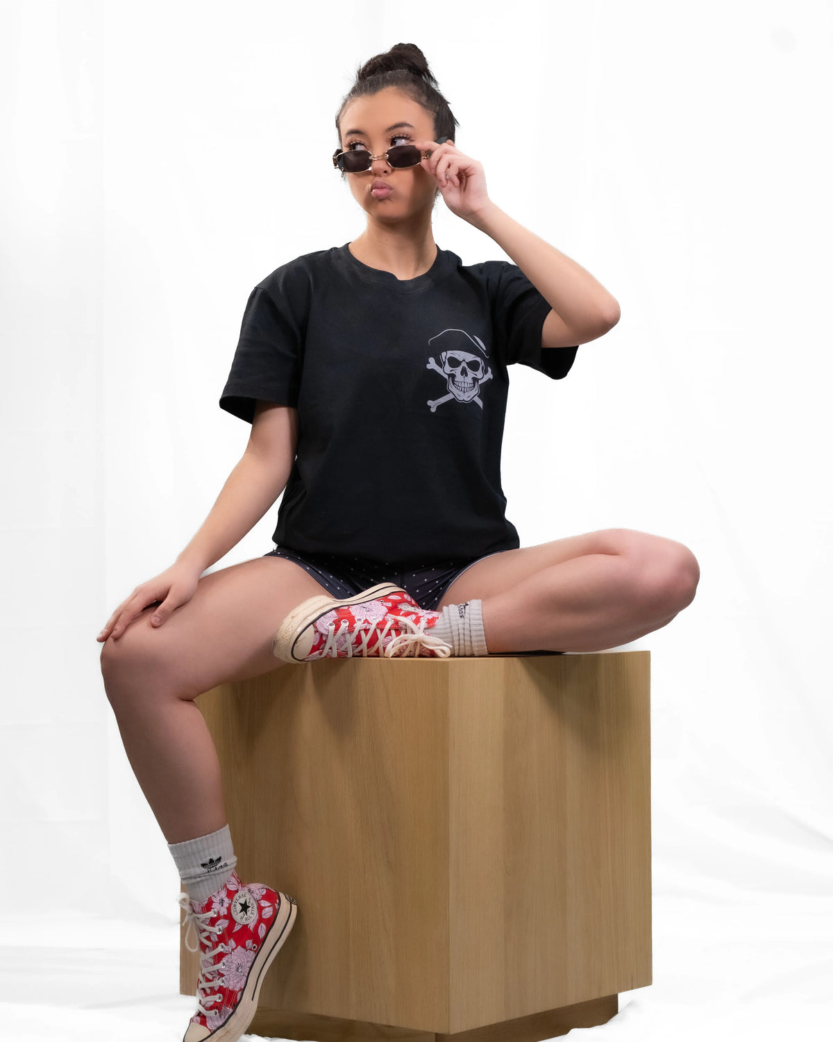 A model sits cross-legged on a wooden block against a white studio background, wearing a black t-shirt with a white skull graphic, patterned shorts, and red-and-white floral high-top sneakers.
