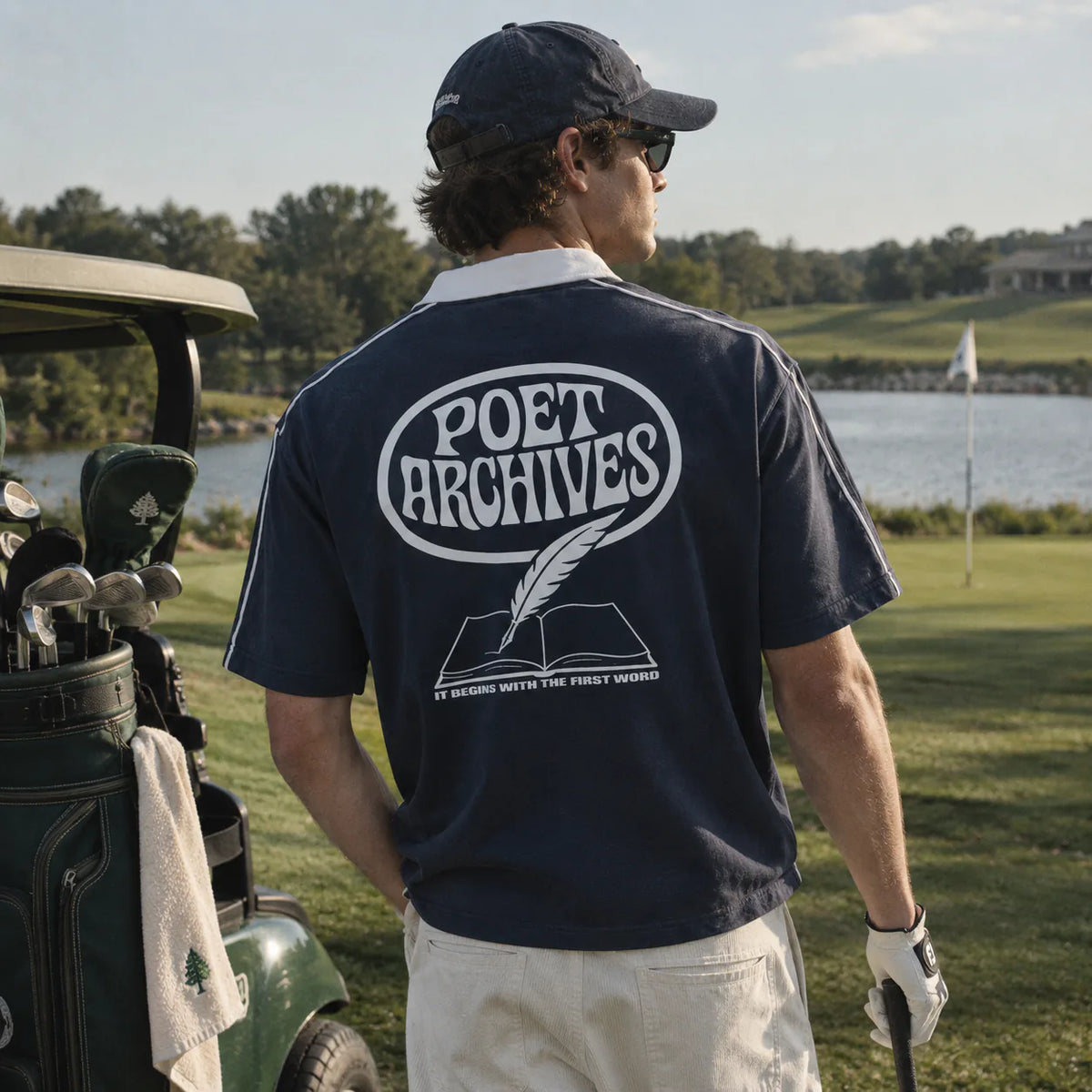 A man on a golf course wearing a navy blue t-shirt with a large white 'POET ARCHIVES' graphic on the back, paired with a navy cap and white pants.