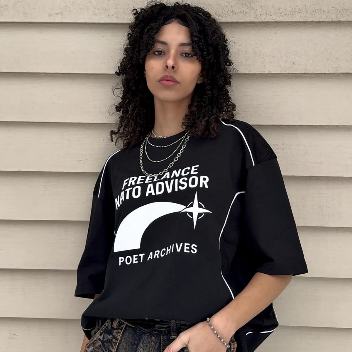 A woman with curly dark hair stands against a beige siding wall wearing a black oversized graphic T-shirt with white text reading 'FREELANCE NATO ADVISOR' and 'POET ARCHIVES,' plus a white curved graphic on the front, paired with dark denim jeans.