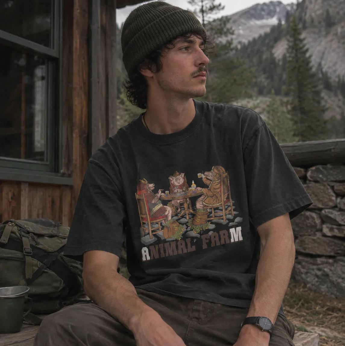 A man wearing a dark charcoal graphic t-shirt with 'ANIMAL FARM' printed on the front, sitting outdoors on a rustic wooden porch with mountains and pine trees in the background.