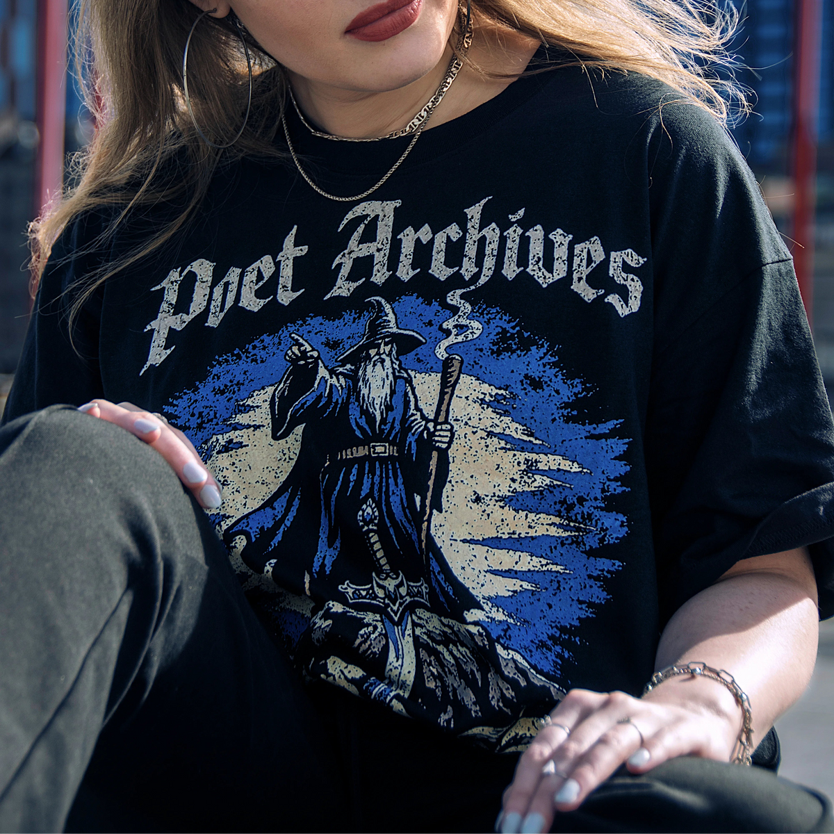A woman wearing a black graphic t-shirt with 'Poet Archives' printed in white gothic lettering above a blue wizard illustration, photographed outdoors against a blurred blue sky and structure background.