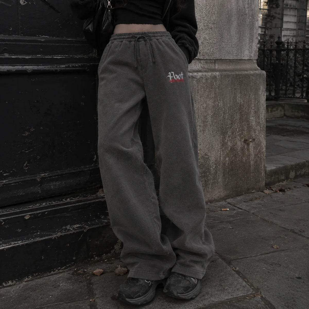 A person wearing loose dark gray sweatpants with a small embroidered logo on the left thigh, standing on a city sidewalk next to a stone pillar.