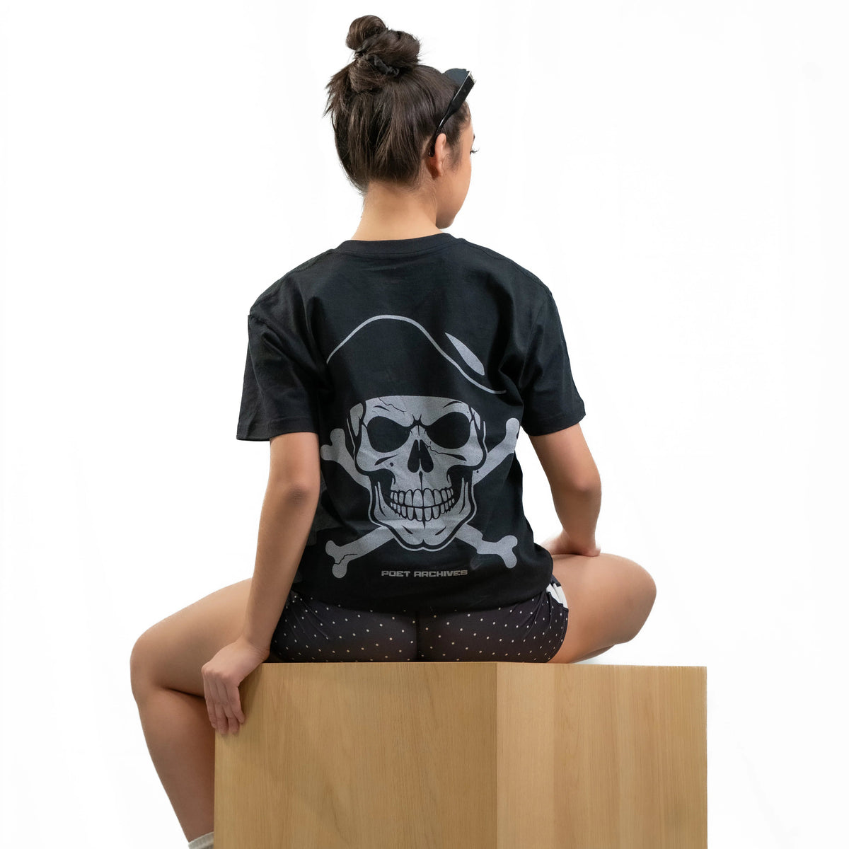 A woman sits on a wooden block from the back, wearing a black t-shirt with a large white pirate skull and crossbones graphic on the back. The shirt features the text 'POET ARCHIVES" below the graphic.