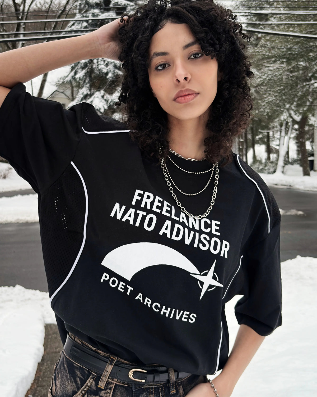 A woman wearing a black graphic t-shirt with white text reading 'FREELANCE NATO ADVISOR' and 'POET ARCHIVES' in a snowy outdoor setting.