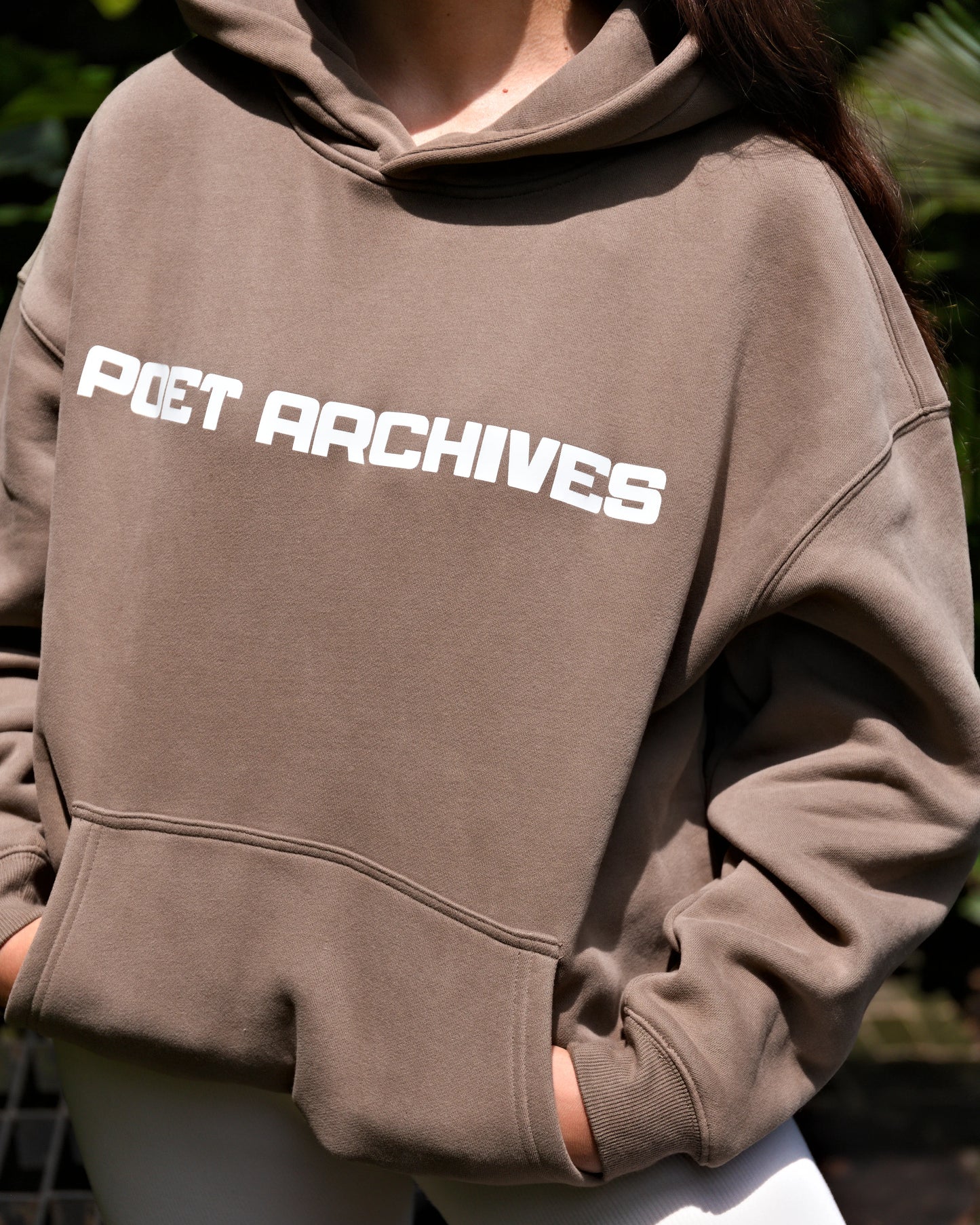 Brown hoodie with 'POET ARCHIVES' text worn by a person, blurred background