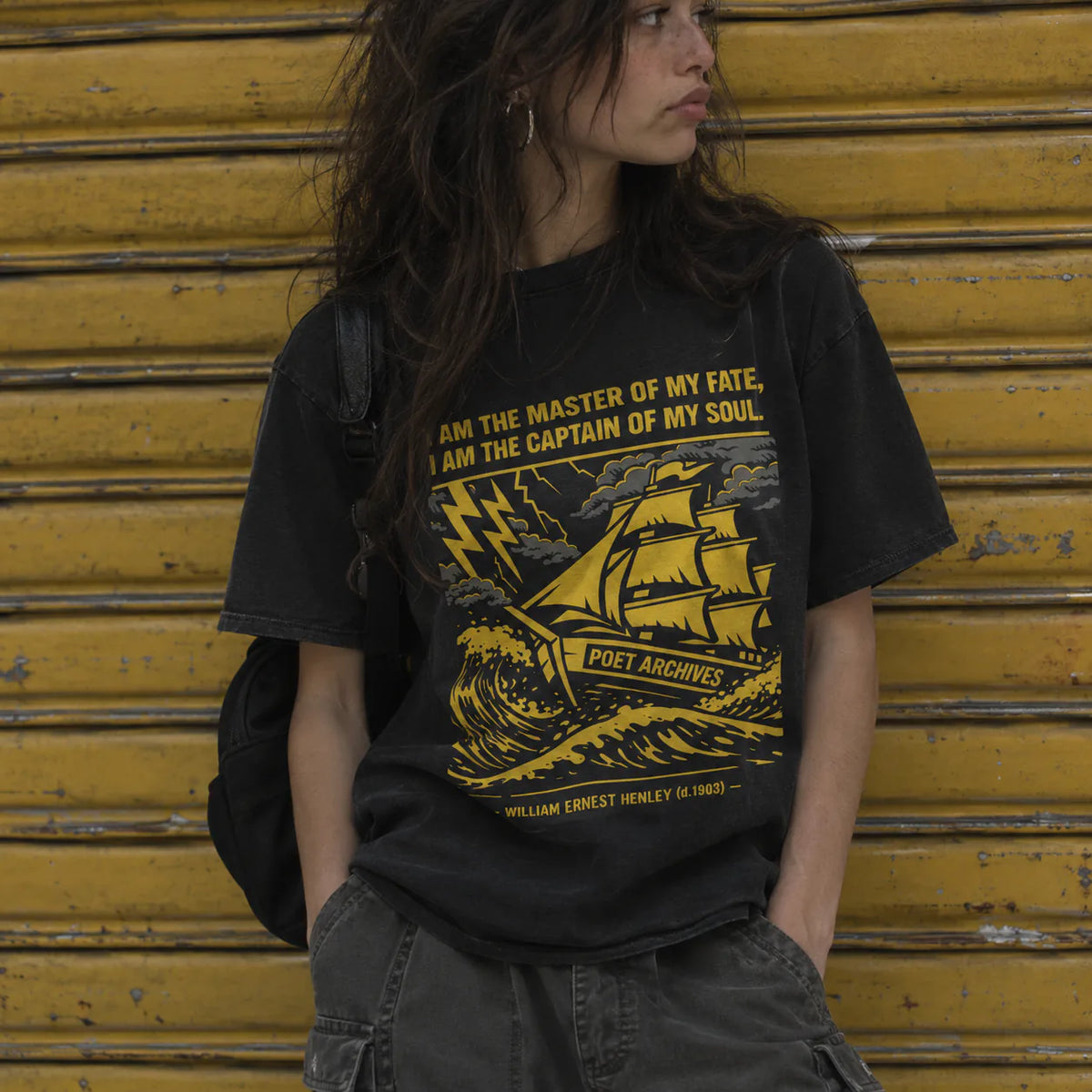 A woman wearing a black graphic t-shirt with a yellow print of a sailing ship and the text 'William Ernest Henry (1869) - I AM THE MASTER OF MY FATE, I AM THE CAPTAIN OF MY SOUL' posed in front of a yellow metal shutter.