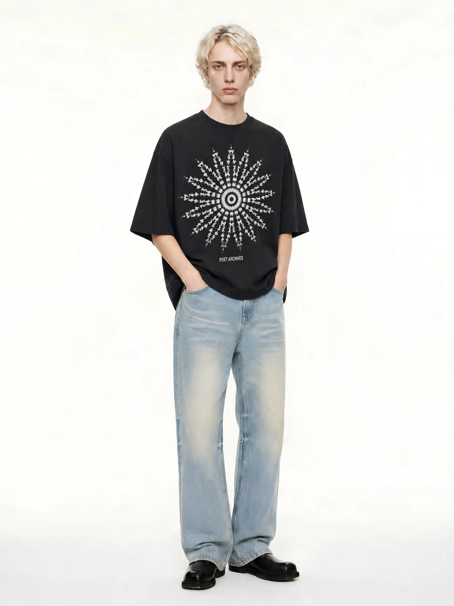 A model stands against a plain white studio background wearing a black oversized T-shirt with a large white geometric starburst graphic on the front, light-wash blue jeans, and black shoes.