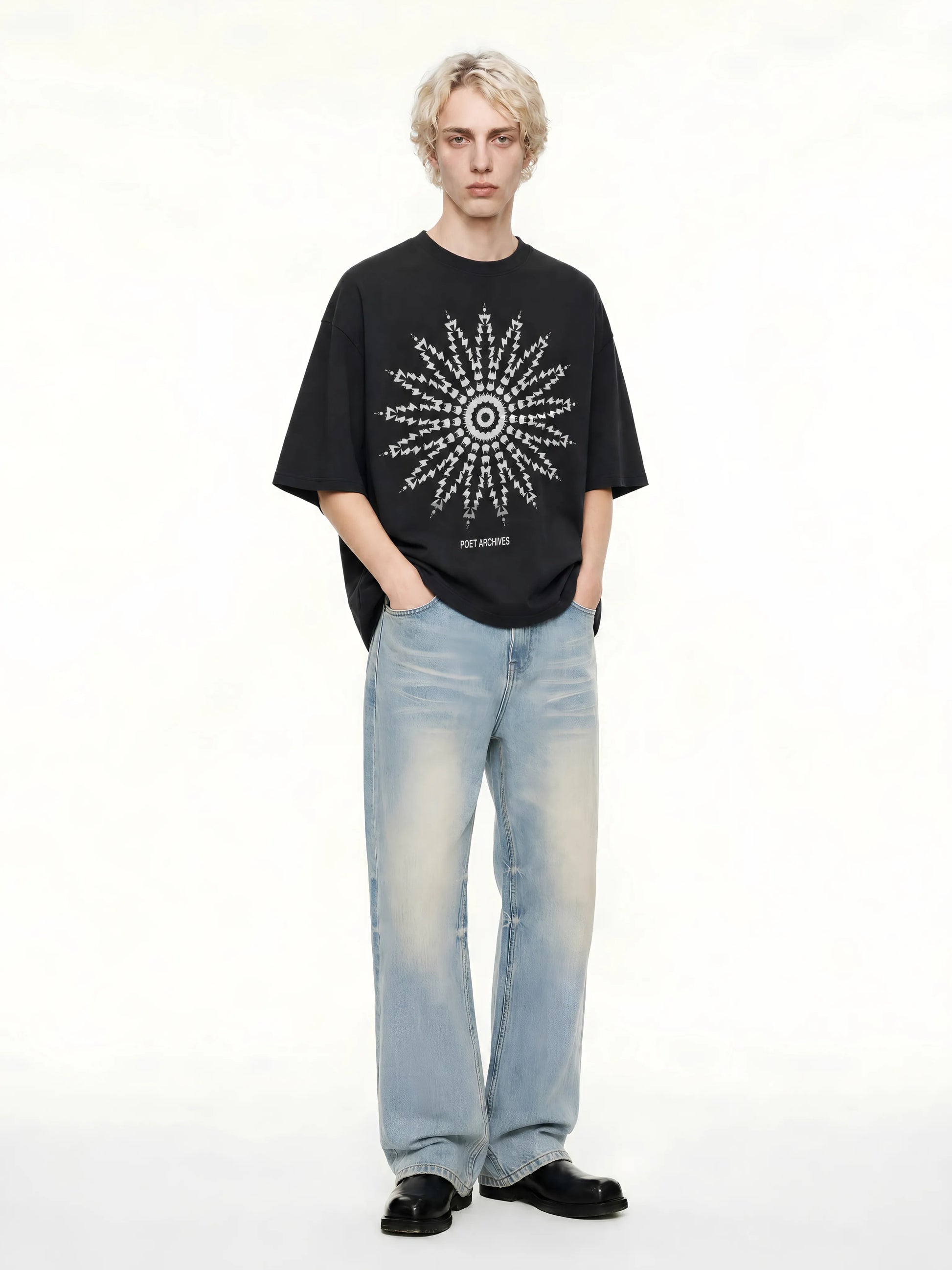 A model stands against a plain white studio background wearing a black oversized T-shirt with a large white geometric starburst graphic on the front, light-wash blue jeans, and black shoes.