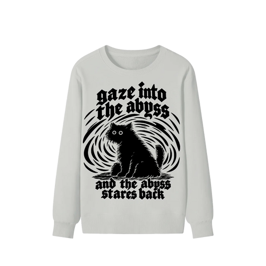 A light gray long-sleeve crewneck sweatshirt featuring a black graphic print of a cat sitting in front of a swirling vortex, with the gothic text 'gaze into the abyss and the abyss stares back'.