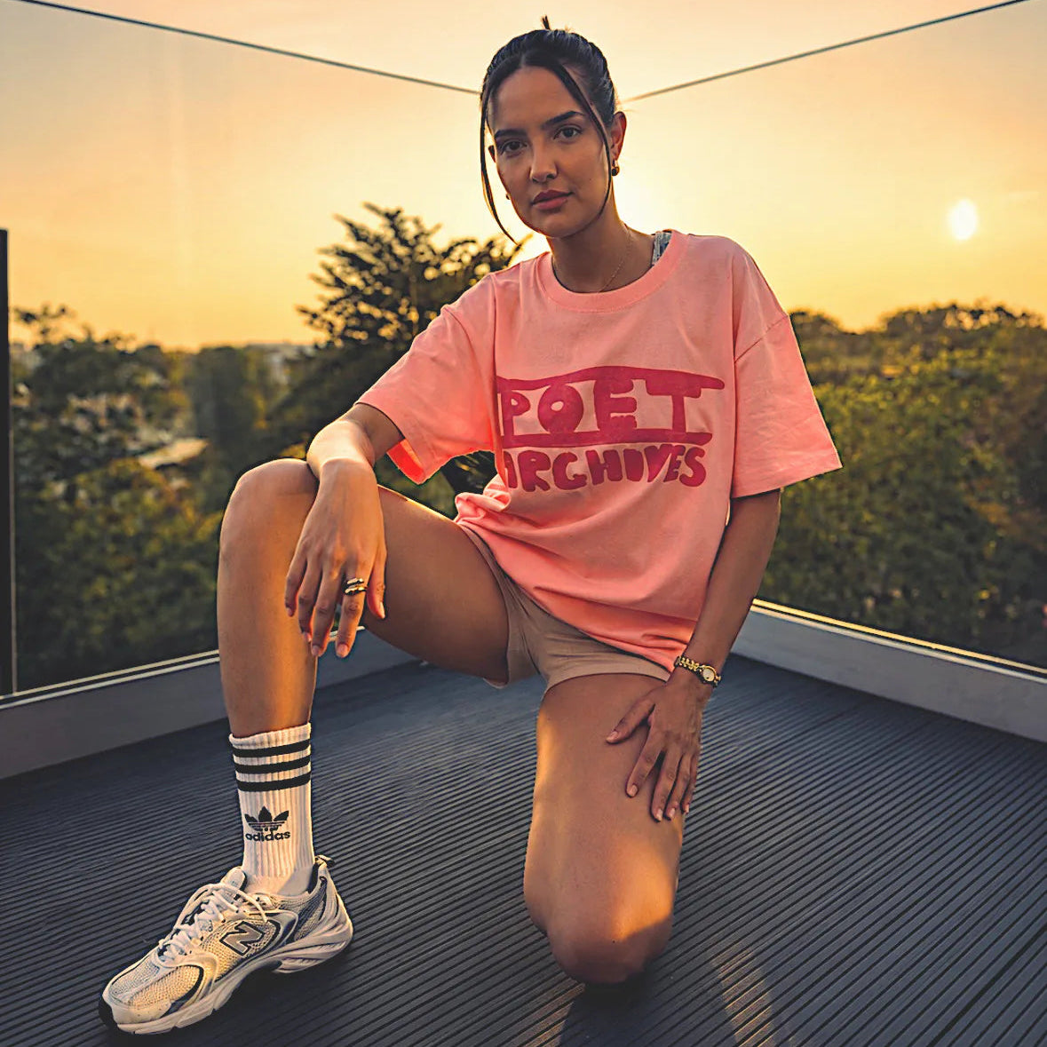 A woman kneels on a rooftop terrace at sunset wearing a pink oversized t-shirt with red graphic text, beige shorts, white socks, and white sneakers.