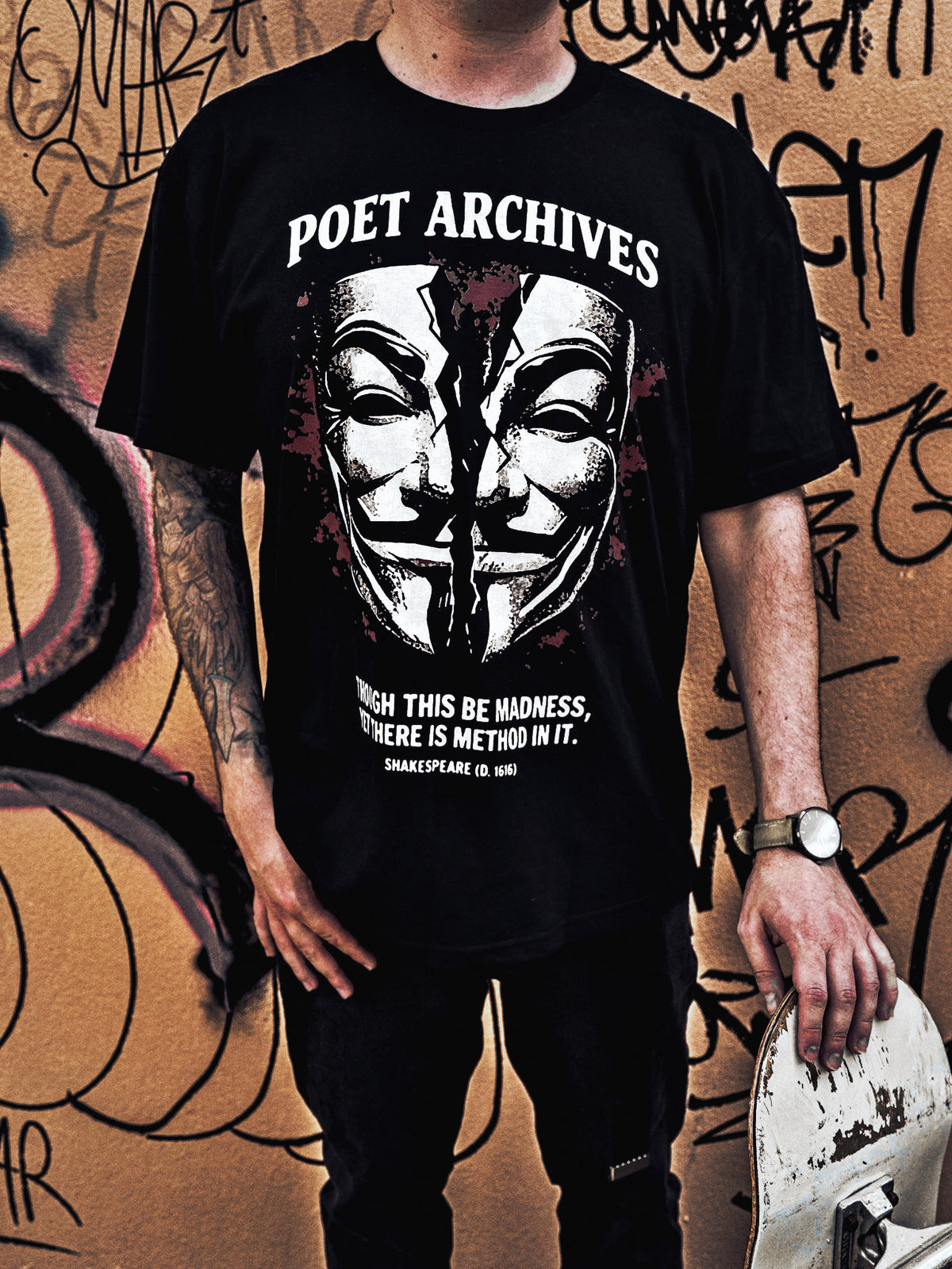 A person wearing a black graphic T-shirt with a large white and red print reading 'POET ARCHIVES' and a masked face design, standing against a graffiti-covered wall and holding a skateboard.