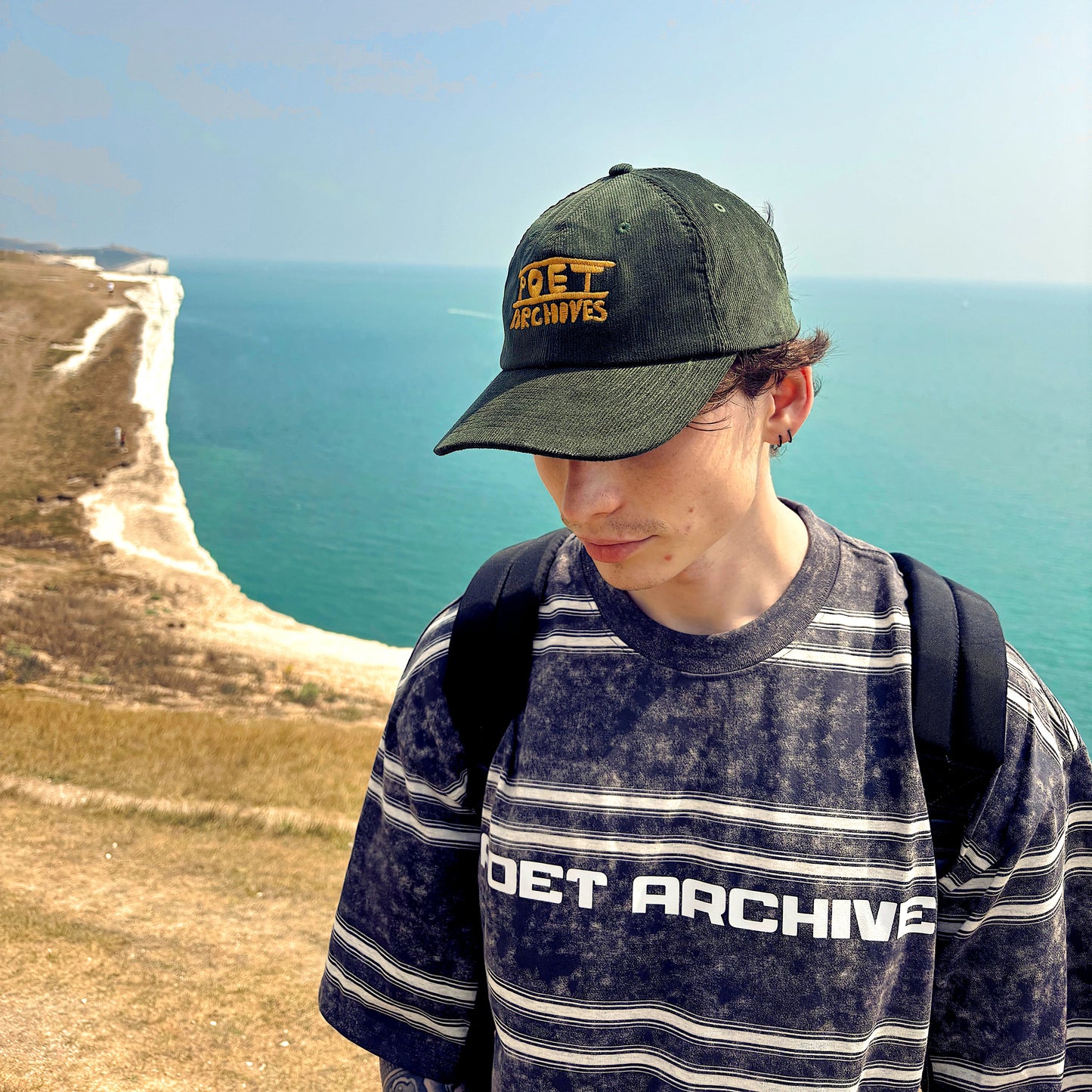 A person outdoors by the sea wearing a dark green cap with yellow 'OET ARCHIVE' embroidery and a matching dark striped T-shirt with 'OET ARCHIVE' text across the chest, against a coastal cliff and blue sky background.
