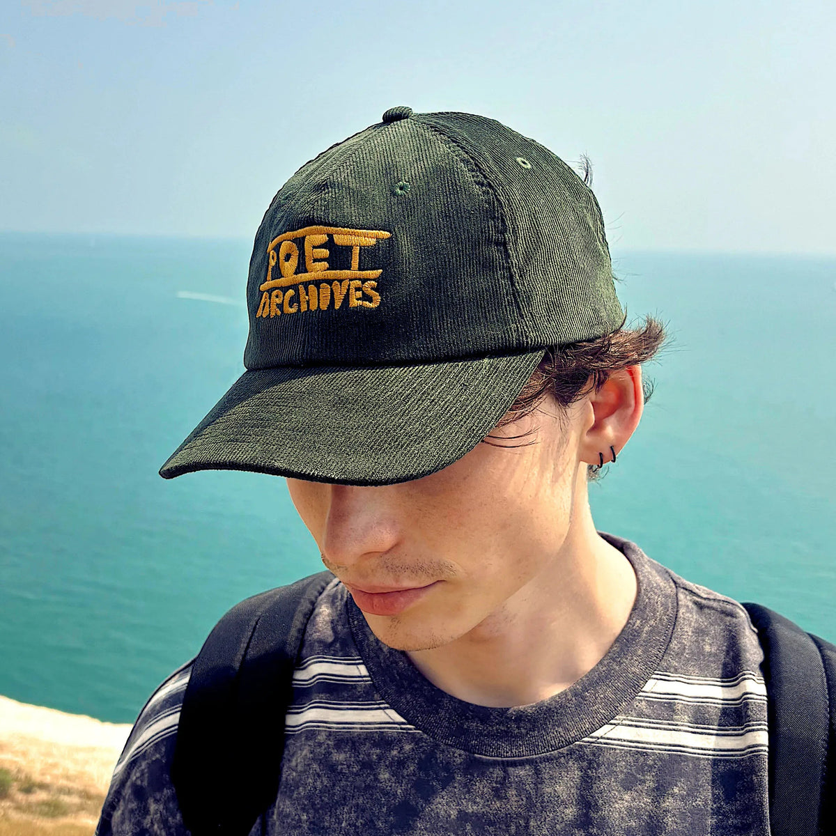 A person standing outdoors on a cliffside overlooking the sea, wearing a dark green cap with yellow 'POET ARCHIVES' text and a dark striped long-sleeve shirt with 'POET ARCHIVES' printed across the chest.