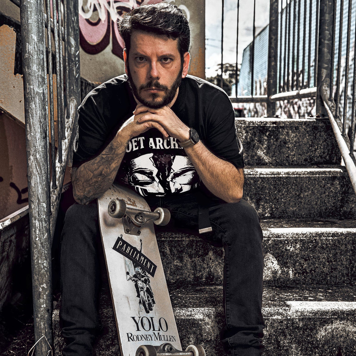 A man with a beard sits on concrete stairs holding a skateboard. He is wearing a black graphic t-shirt, dark jeans, and black canvas sneakers.