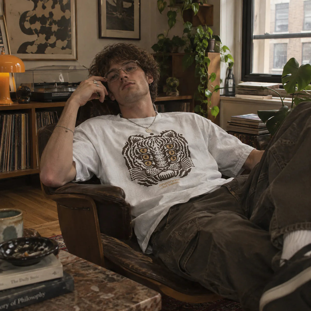 A man reclining in a wooden armchair wearing a white graphic t-shirt with a black and gold tiger print, brown cargo pants, and glasses in a cozy, book-filled room.