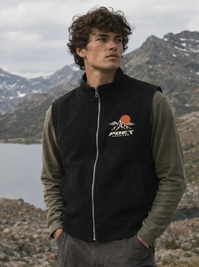 A man wearing a black zip-up fleece vest with a white and orange Poet Archives logo, layered over an olive green long-sleeve shirt, standing in a mountainous outdoor setting.