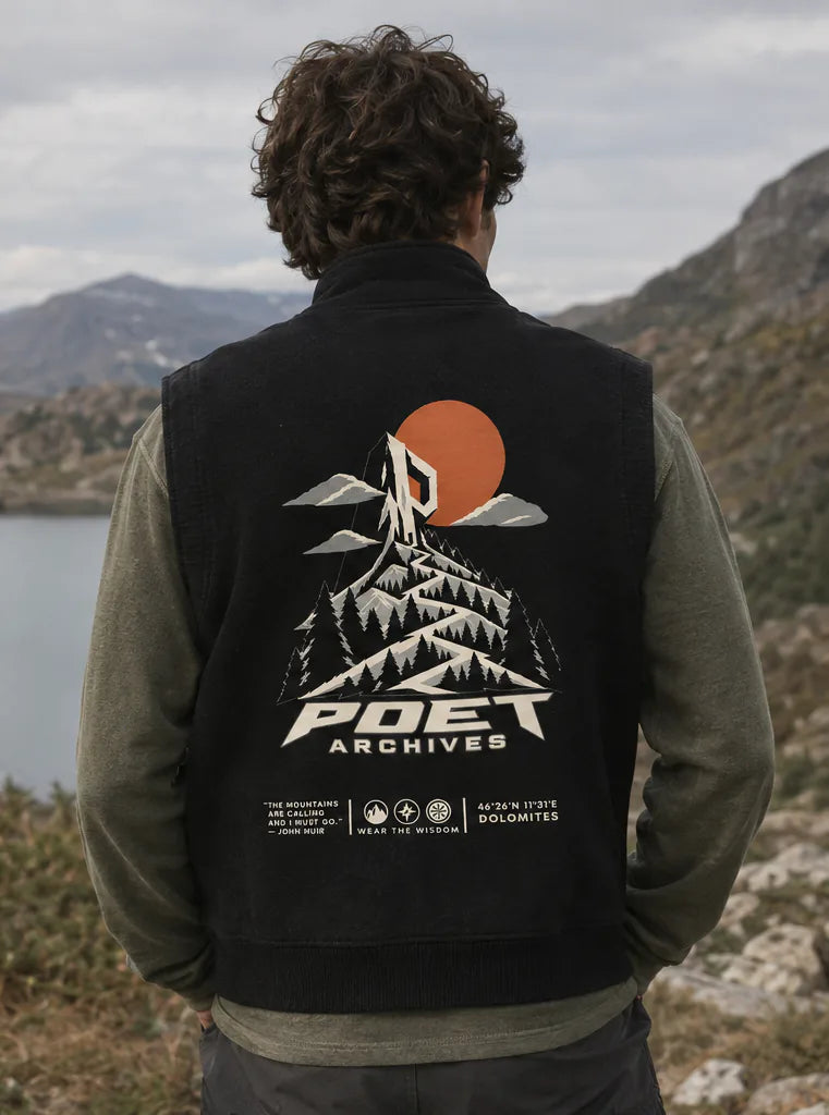 A man viewed from behind wearing a black wool-blend vest with a large graphic print on the back, standing outdoors in front of mountains and a lake. The vest features an orange sun, white mountains, and the text 'POET ARCHIVES'.