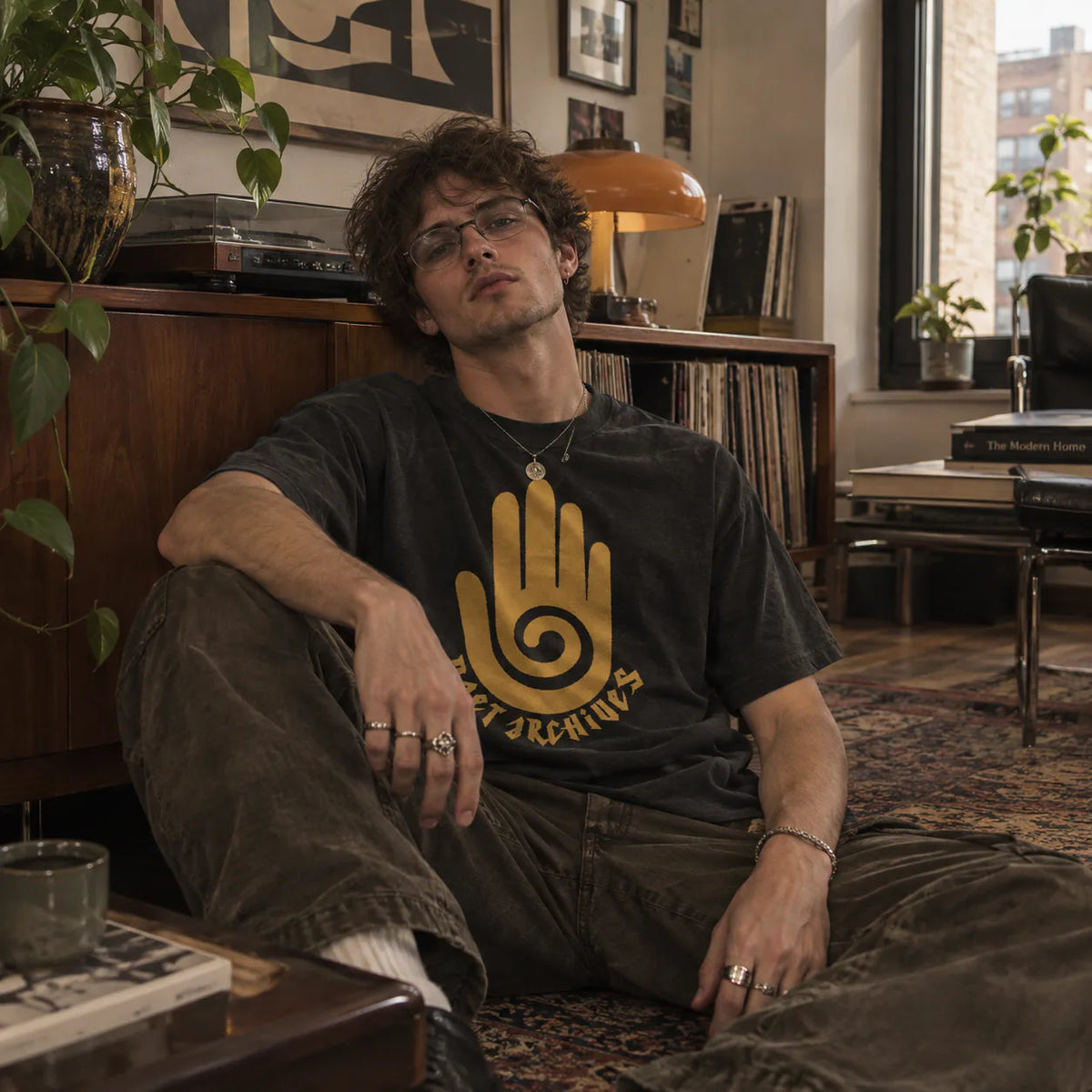 A man wearing a black graphic t-shirt with a gold spiral hand design and the text 'ALL THINGS ARE' sits on a patterned rug in a cozy, vintage-styled room with records and plants.