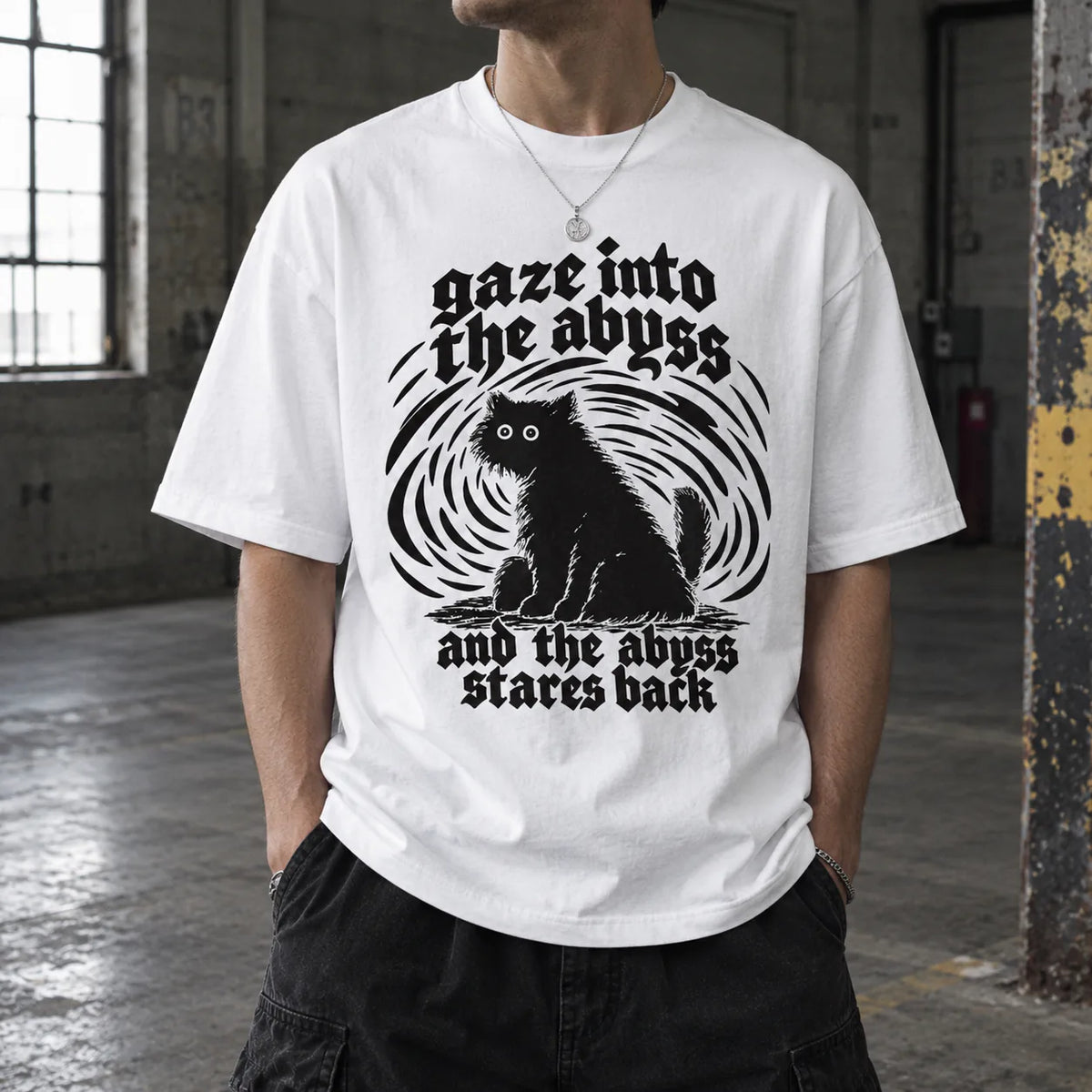 A person wearing a white graphic T-shirt in an industrial loft setting. The shirt features a black circular swirl design with a black cat silhouette in the center and gothic text reading 'gaze into the abyss and the abyss stares back'.